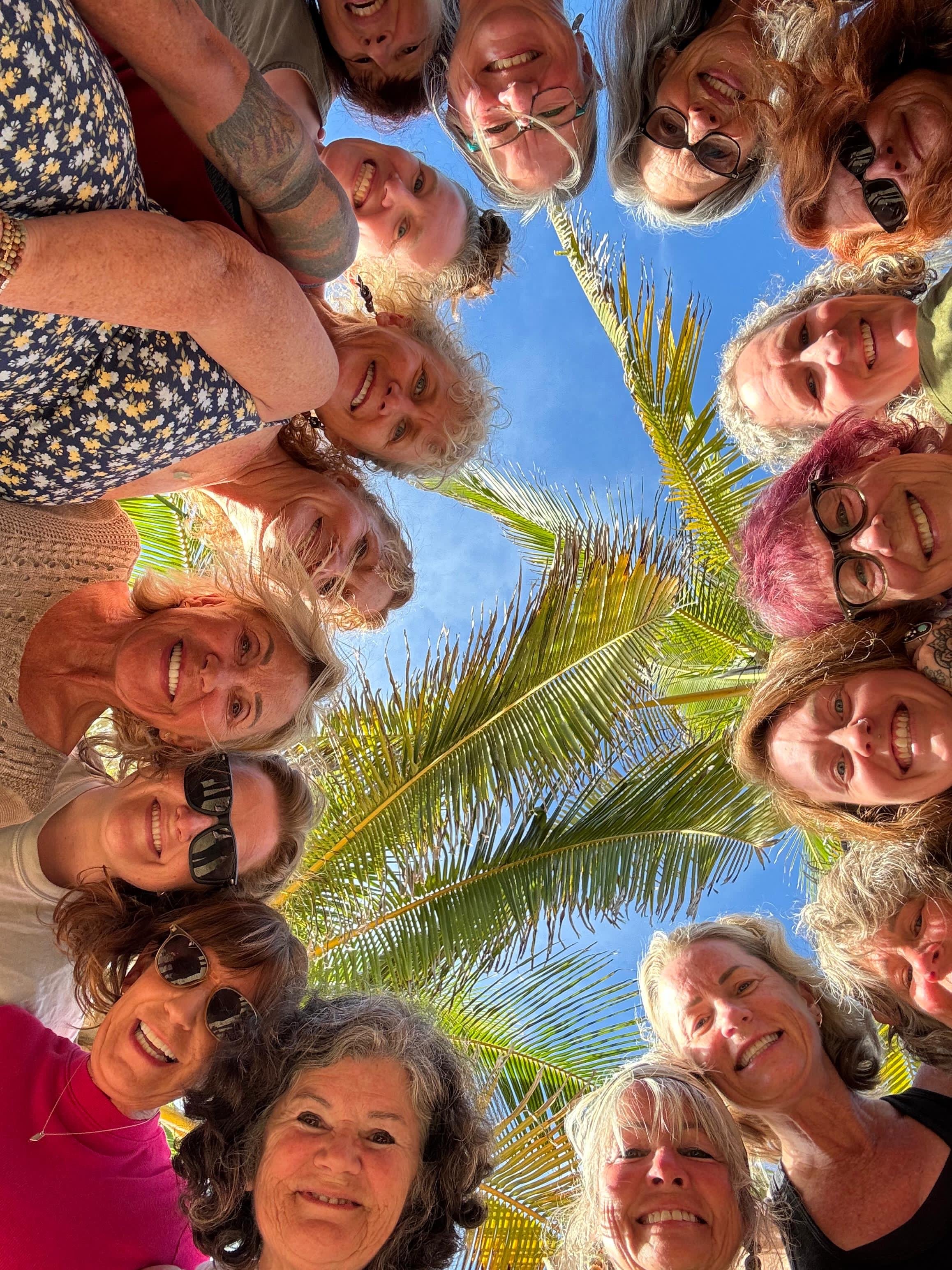 A group of diverse women smiling and looking down into the camera, taken outdoors with clear blue sky and palm leaves visible above them.