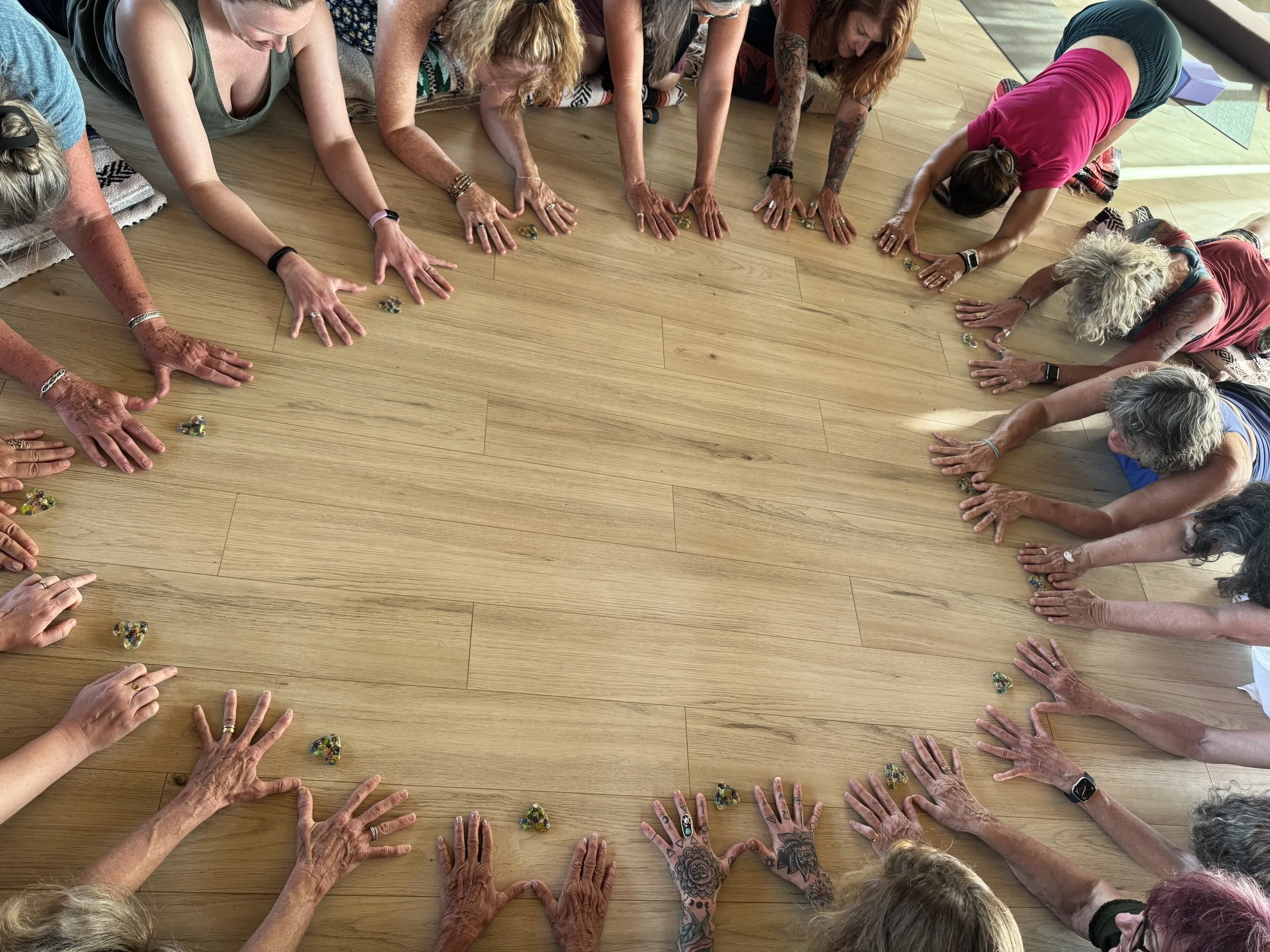 Group of women practicing yoga or a similar activity on a wooden floor, with their hands extended forward, some wearing colorful clothing and visible tattoos.