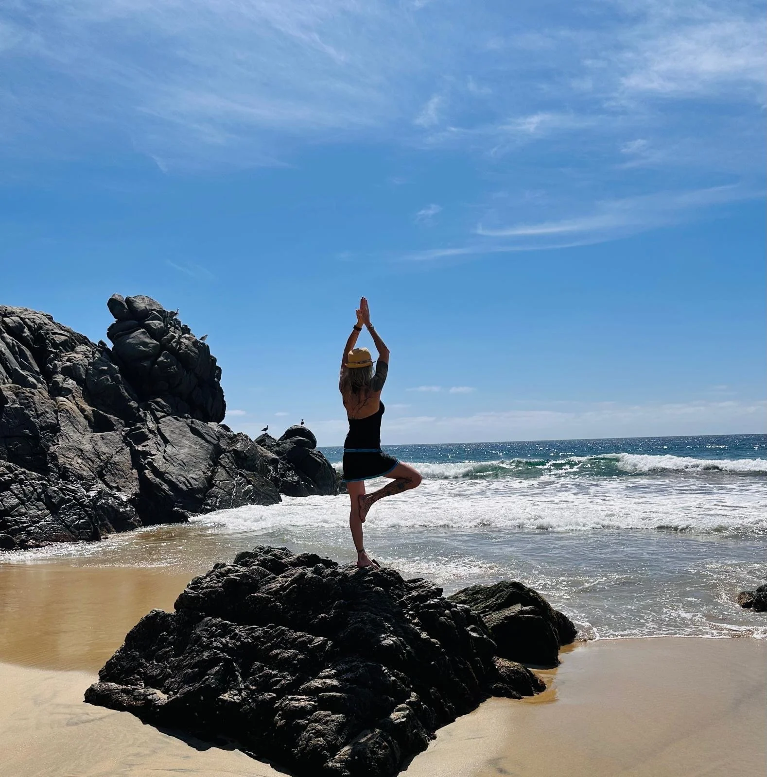 Woman practicing yoga on rocks at the beach during daytime under a blue sky with clouds.