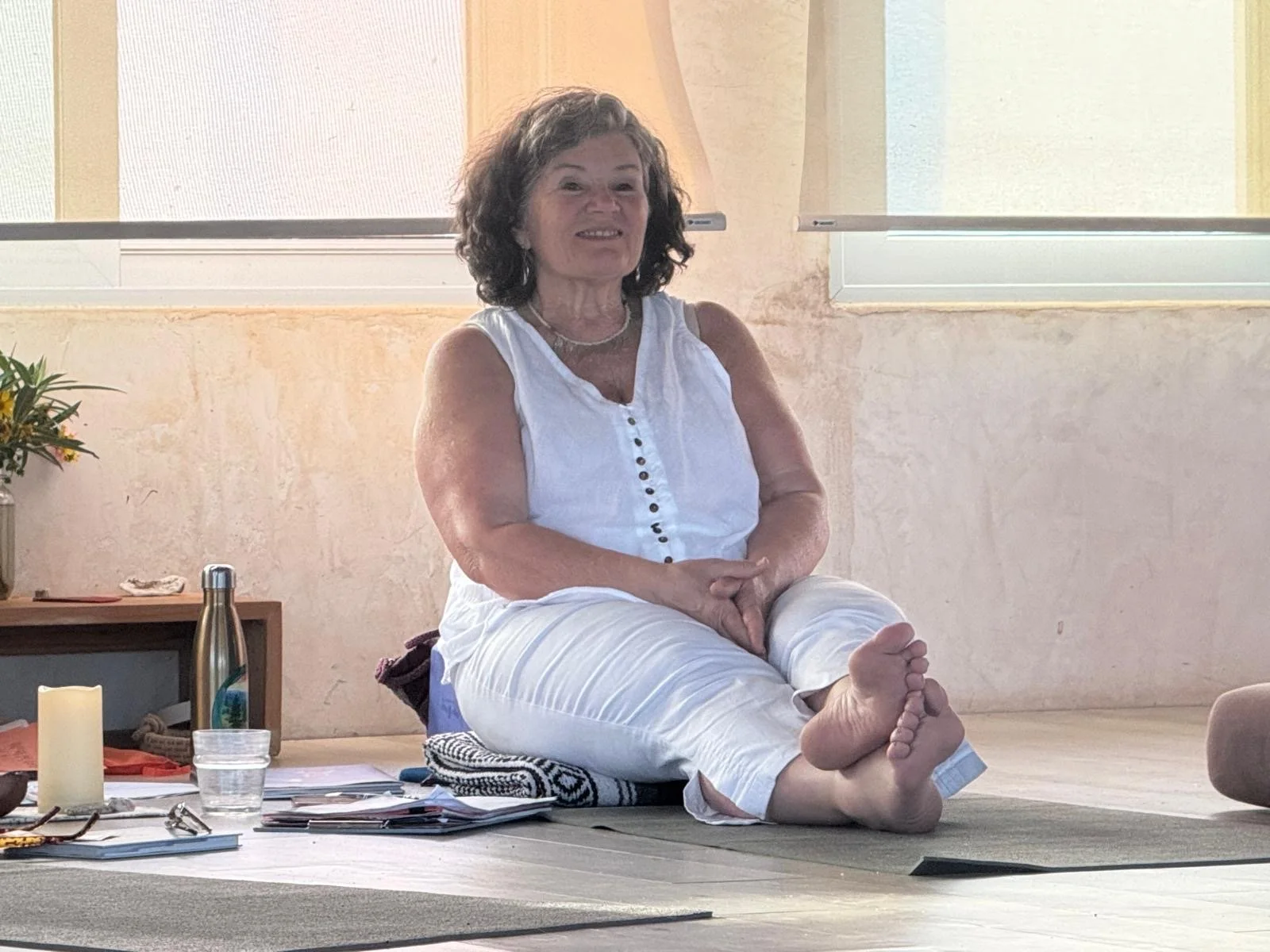 A woman practicing yoga indoors, sitting cross-legged on a cushion, with her feet resting on her knees, smiling at the camera, with a table and window behind her.