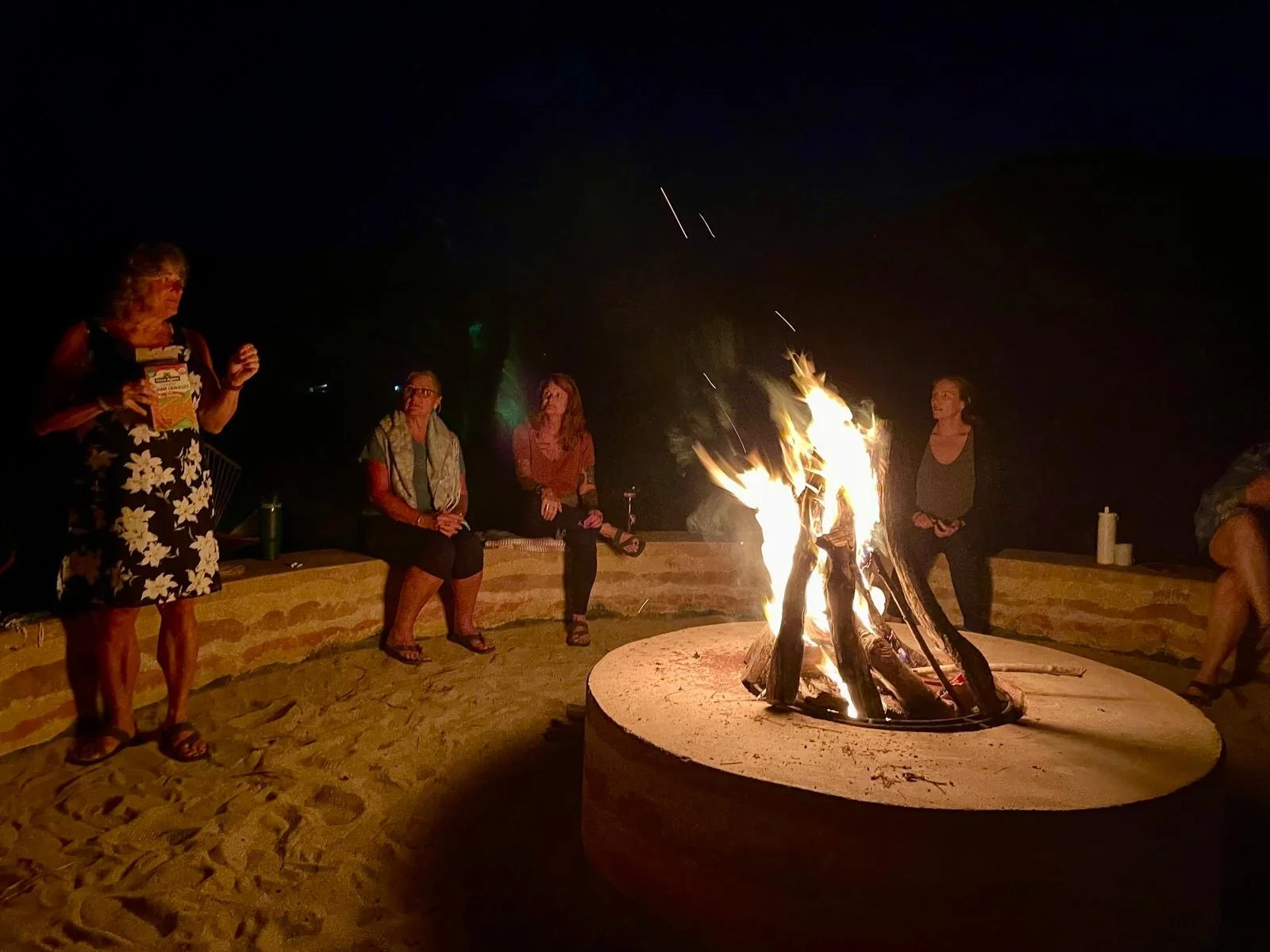 Group of people sitting around a campfire at night, some seated on a low wall, with one woman standing, holding a snack bag, while others watch the fire and chat.