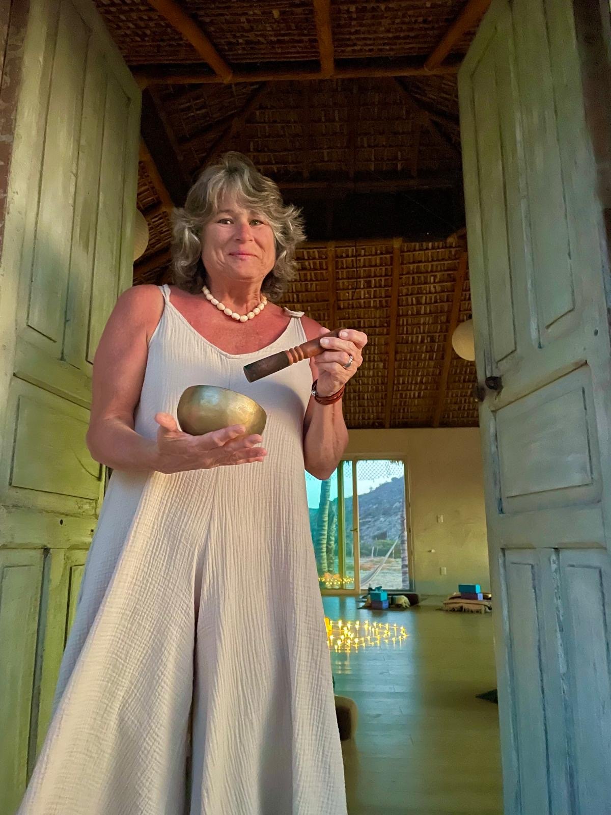 A woman holding a singing bowl and a wooden mallet, standing at the entrance of a room with open doors, inside a wooden structure with a high, thatched ceiling and a view of nature outside.