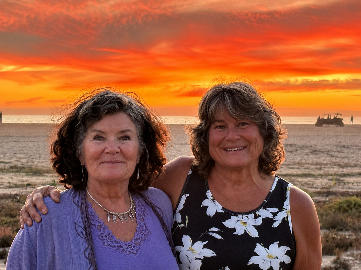 Two women smiling on a beach at sunset, with colorful clouds and the ocean in the background.