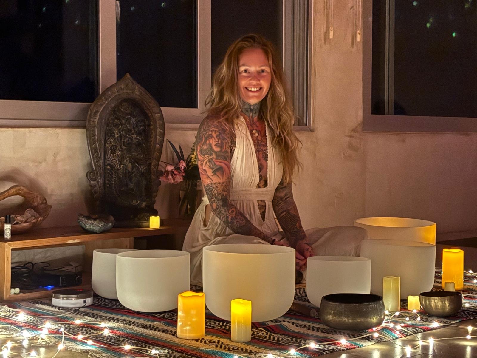 A smiling woman with long, wavy, red hair and tattoos on her arms and chest, sitting on a colorful patterned rug surrounded by candles and singing bowls in a cozy indoor setting at night.