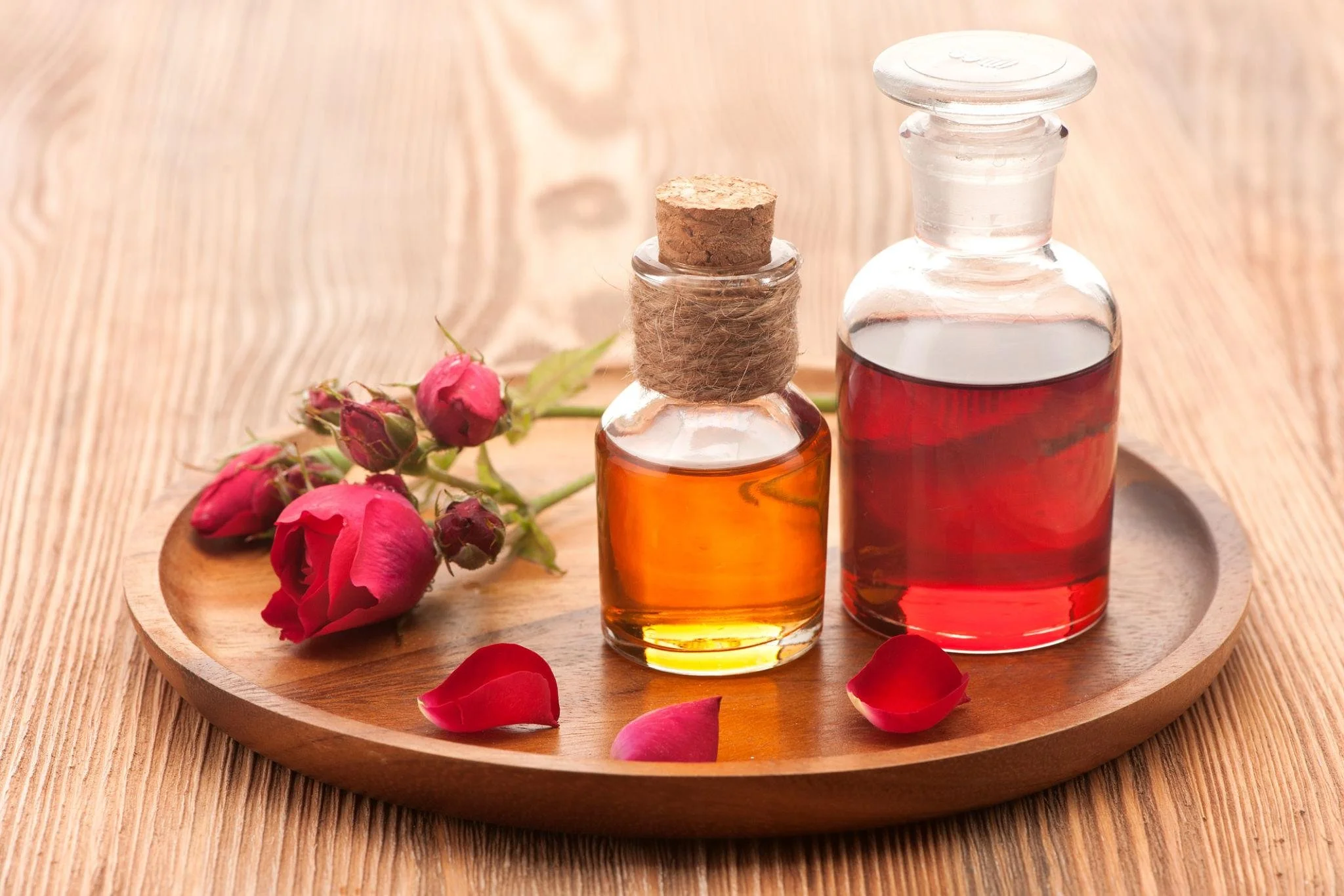 Two small glass bottles containing red and orange liquids, placed on a round wooden tray with pink rosebuds and scattered rose petals, on a wooden surface.