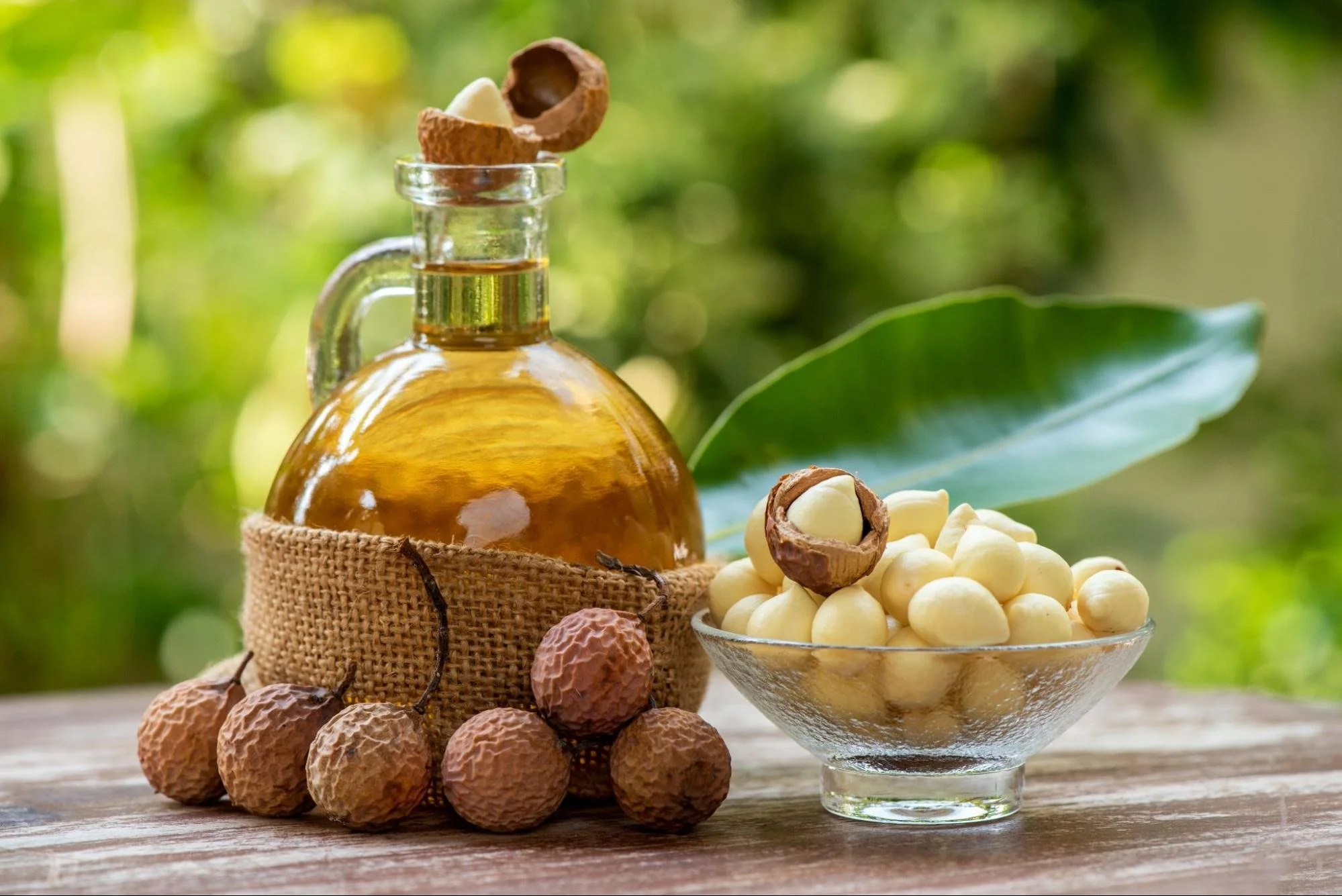 A glass bowl filled with white macadamia nuts, a small jar of macadamia oil, and a sack of whole macadamia nuts, with some opened shells and nuts scattered around on a wooden surface, against a blurred green outdoor background.