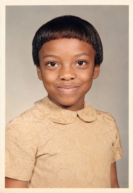 A young boy with short black hair and a slight smile, wearing a beige patterned shirt against a neutral background.