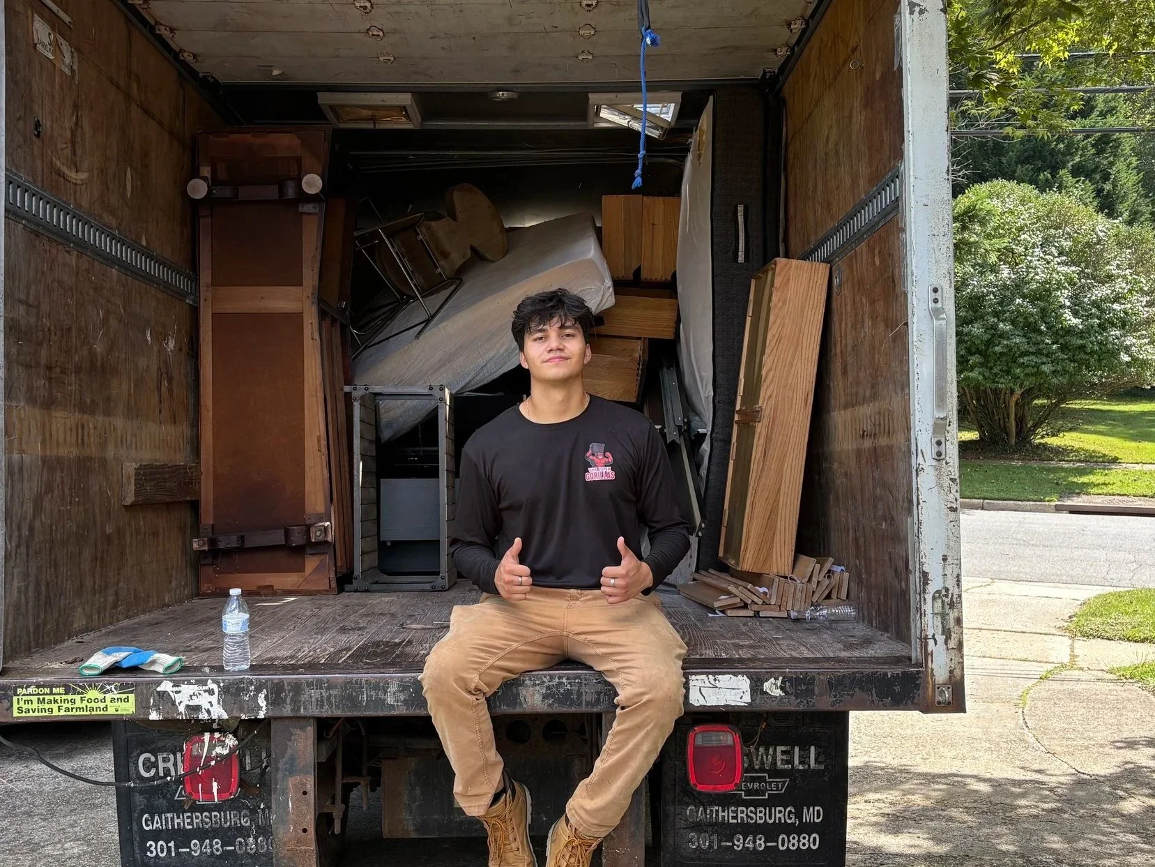 Young man sitting on the back of a moving truck with furniture inside, giving a thumbs-up gesture. It is daytime, and there is a neighborhood street with trees in the background.