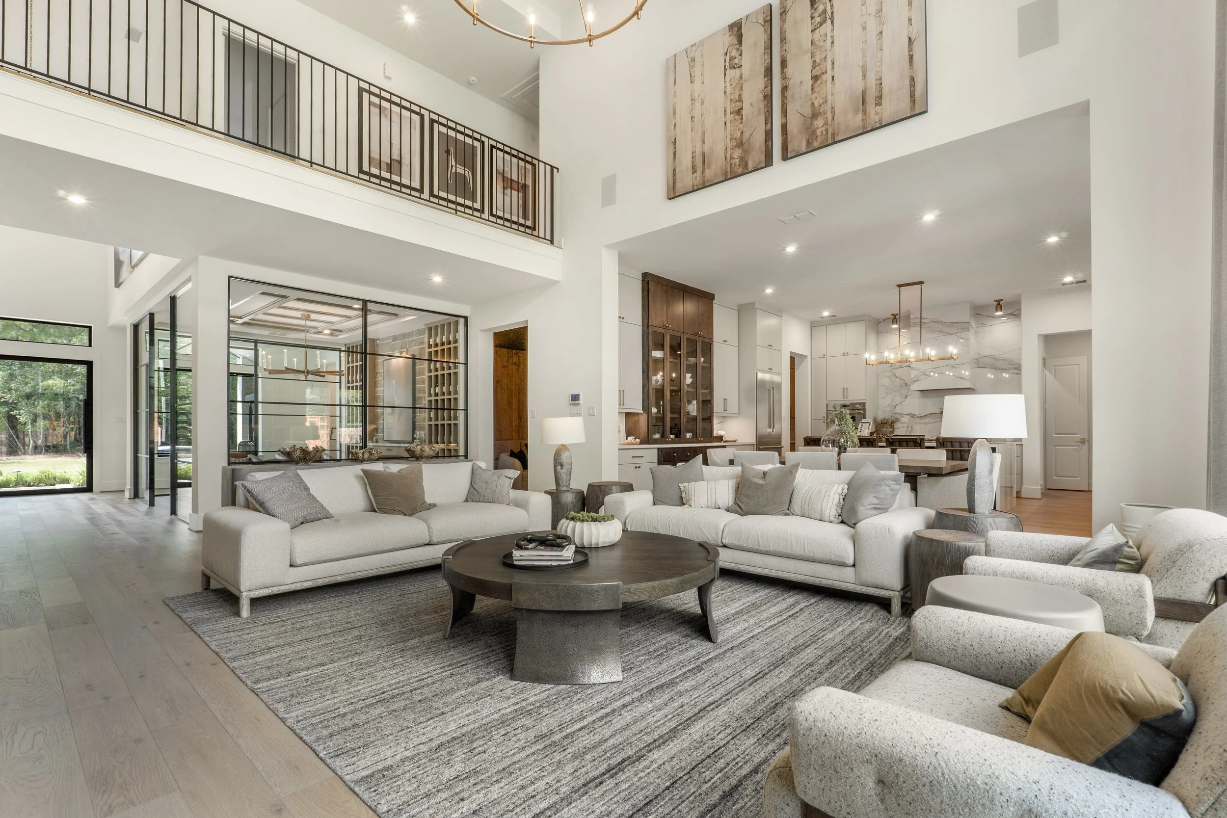 Living room in a modern home with white sofas, gray cushions, a round wooden coffee table, a patterned gray rug, and contemporary decor. The open concept includes a view of the kitchen with white cabinets, a marble backsplash, and a dining area with 