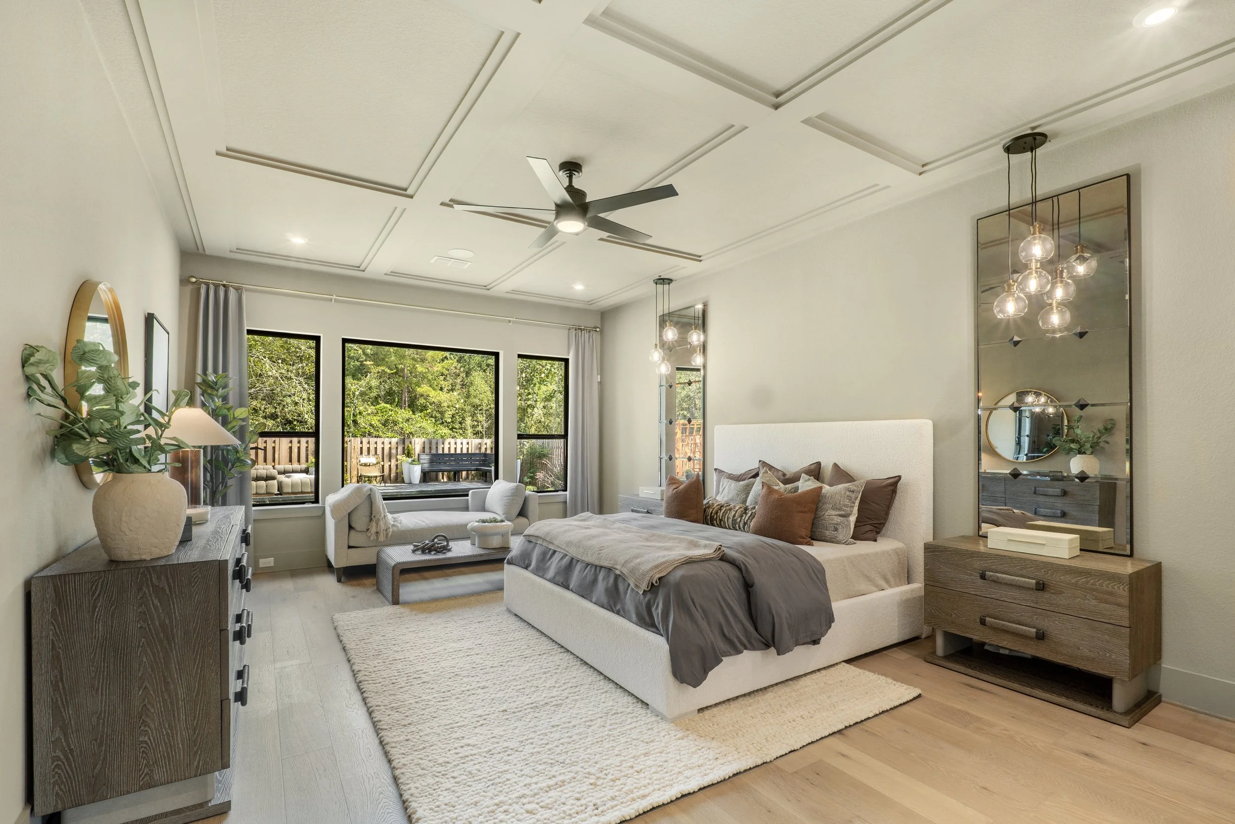 Bright bedroom with large window, white bed with pillows, wooden nightstand, modern chandelier, gray and beige decor, outdoor balcony with seating, wooden flooring, and ceiling fan.