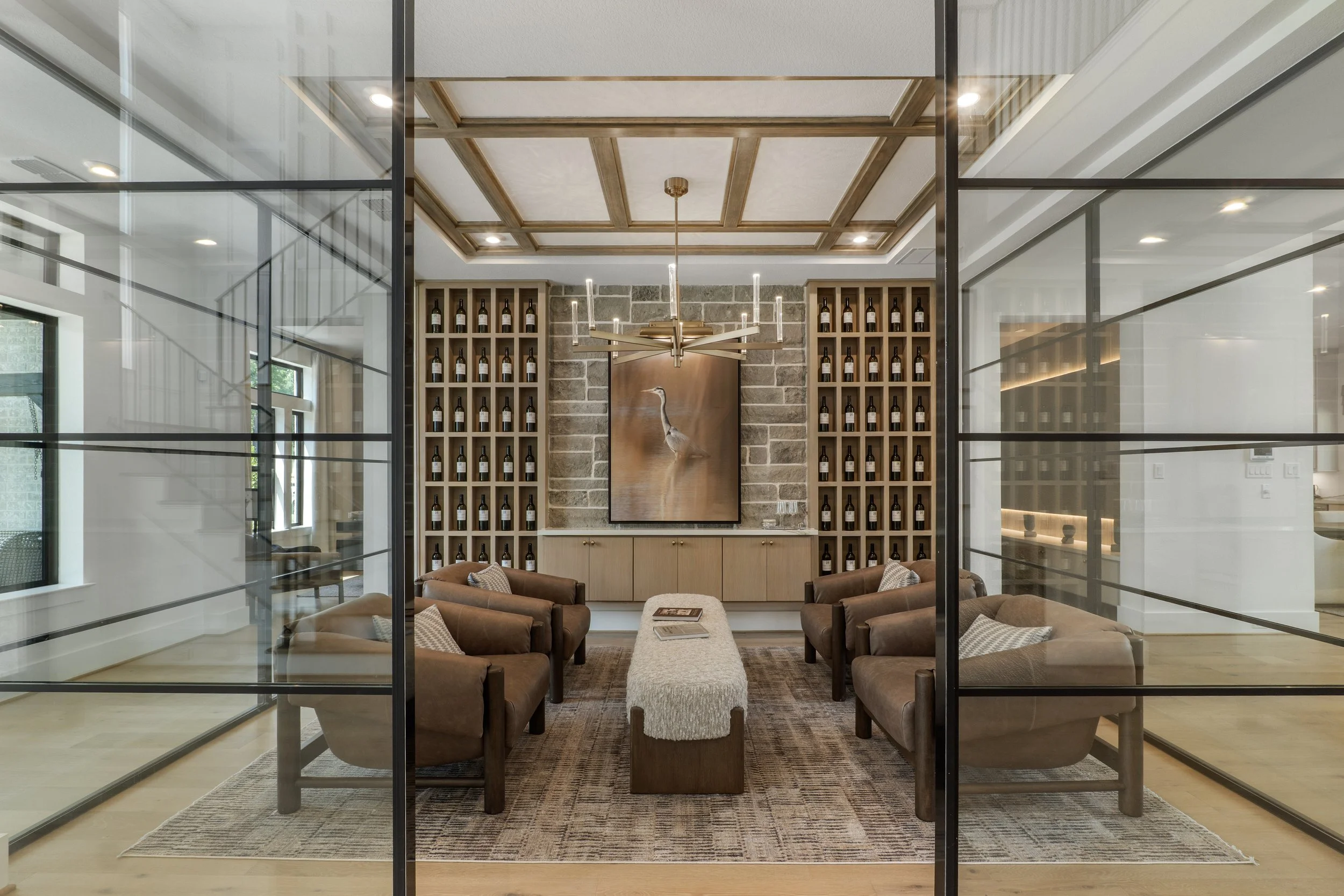 A cozy sitting area with four brown armchairs around a rectangular beige ottoman, a rug underneath, and a wall of wine bottles on wood shelves behind, with a metallic bird painting and a modern chandelier hanging from a coffered ceiling.