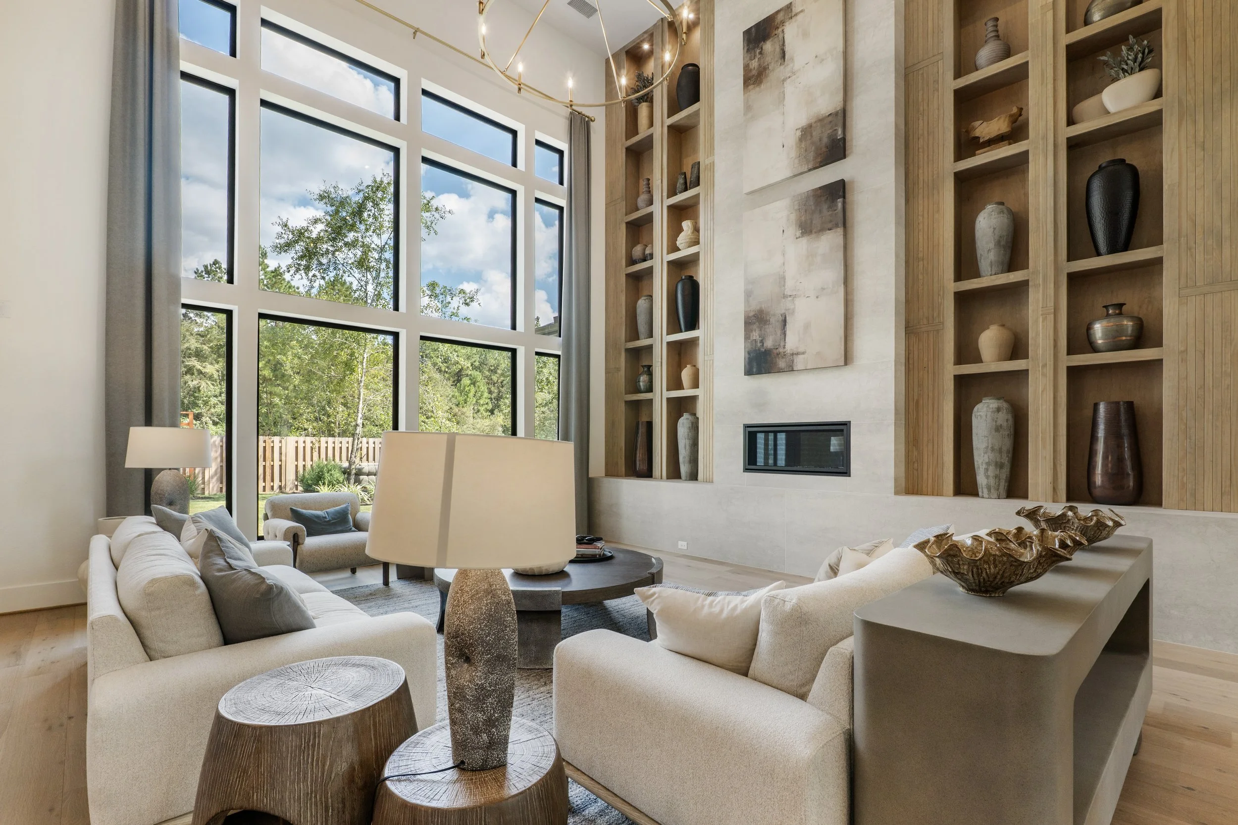 A modern living room with large floor-to-ceiling windows, beige sofas, wooden furniture, and decorative shelves with vases. There's a circular chandelier hanging from the ceiling, a painting on the wall, and a view of the outdoor garden with trees.