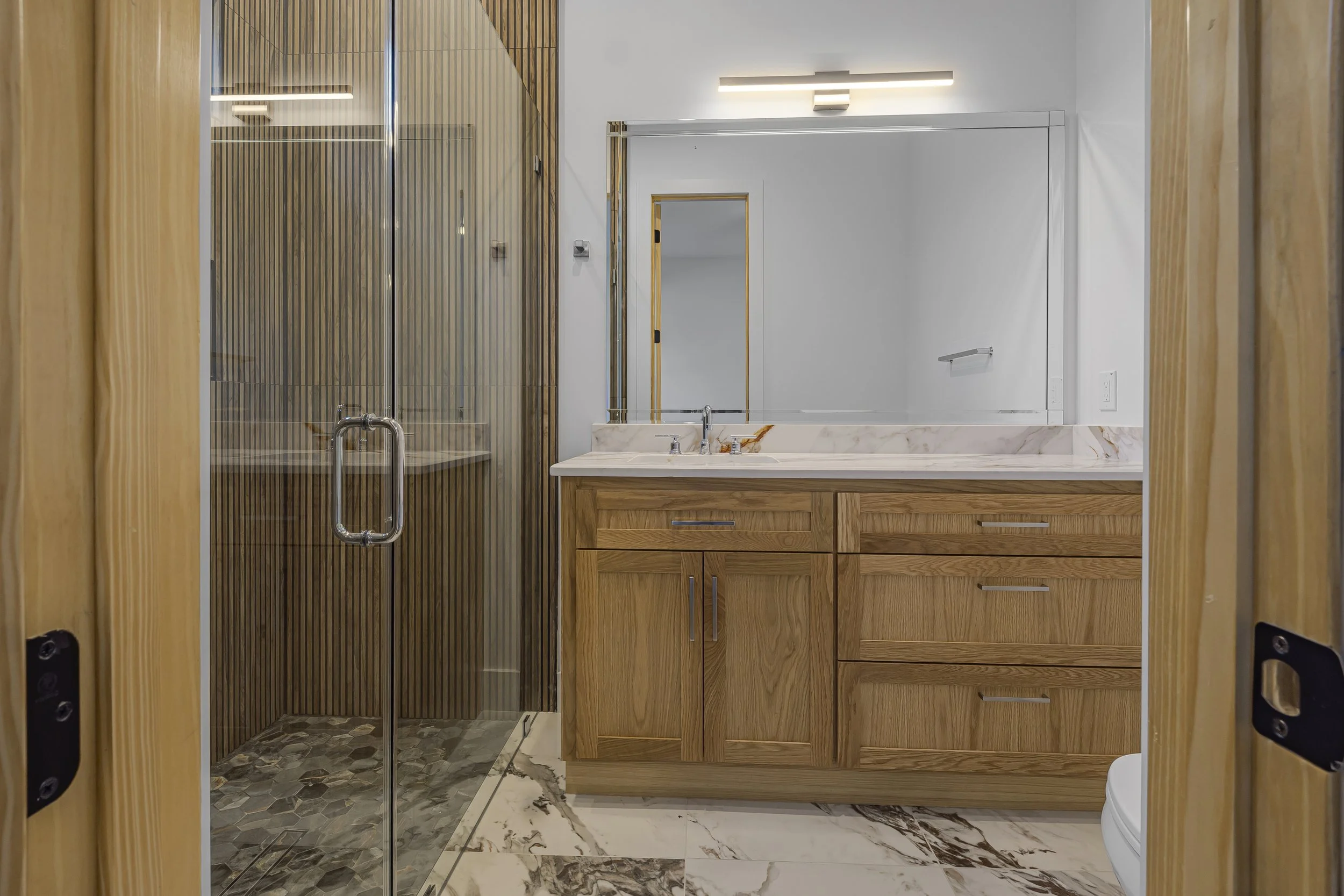 Modern bathroom with a glass-enclosed shower on the left, wooden vanity with a marble countertop and a large mirror above, and marble floor tiles.