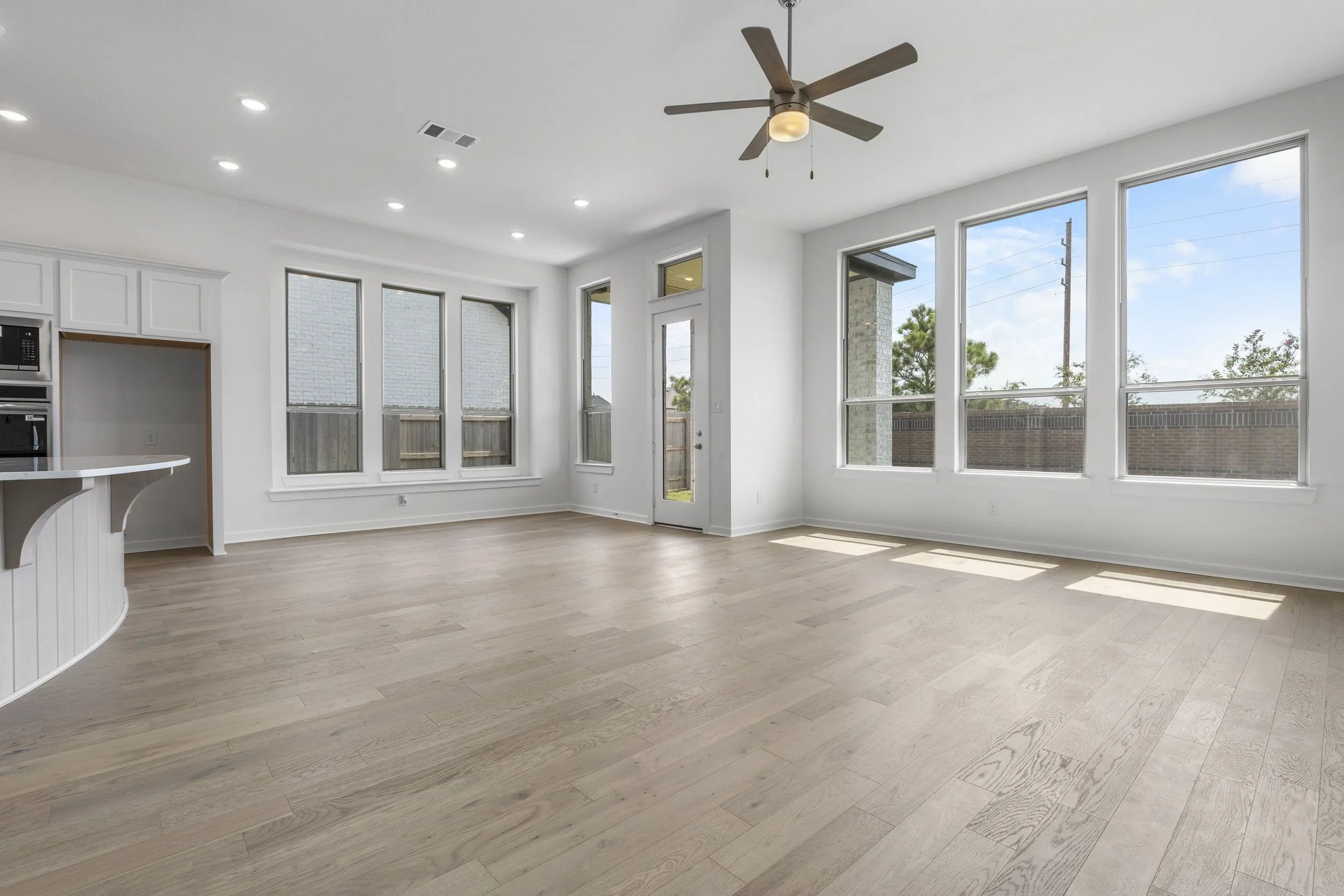 Empty living room with large windows, wooden floor, ceiling fan, and white walls.