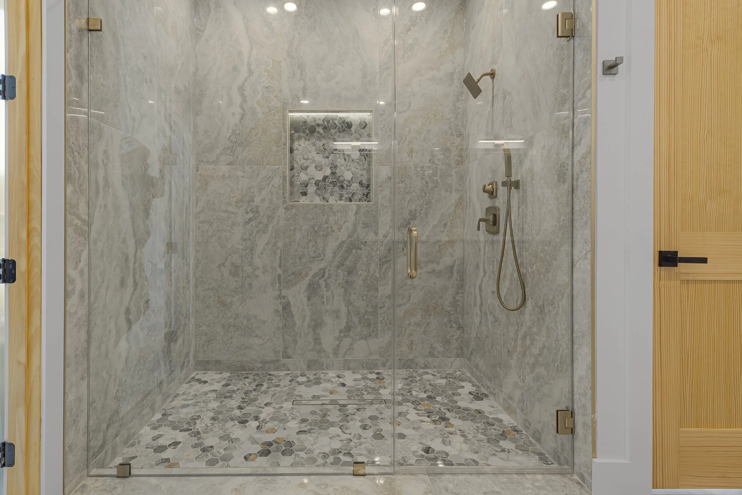 A modern glass shower enclosure with brass hardware, a built-in tile niche, and a mosaic tile floor in a bathroom with beige and gray marble surfaces.