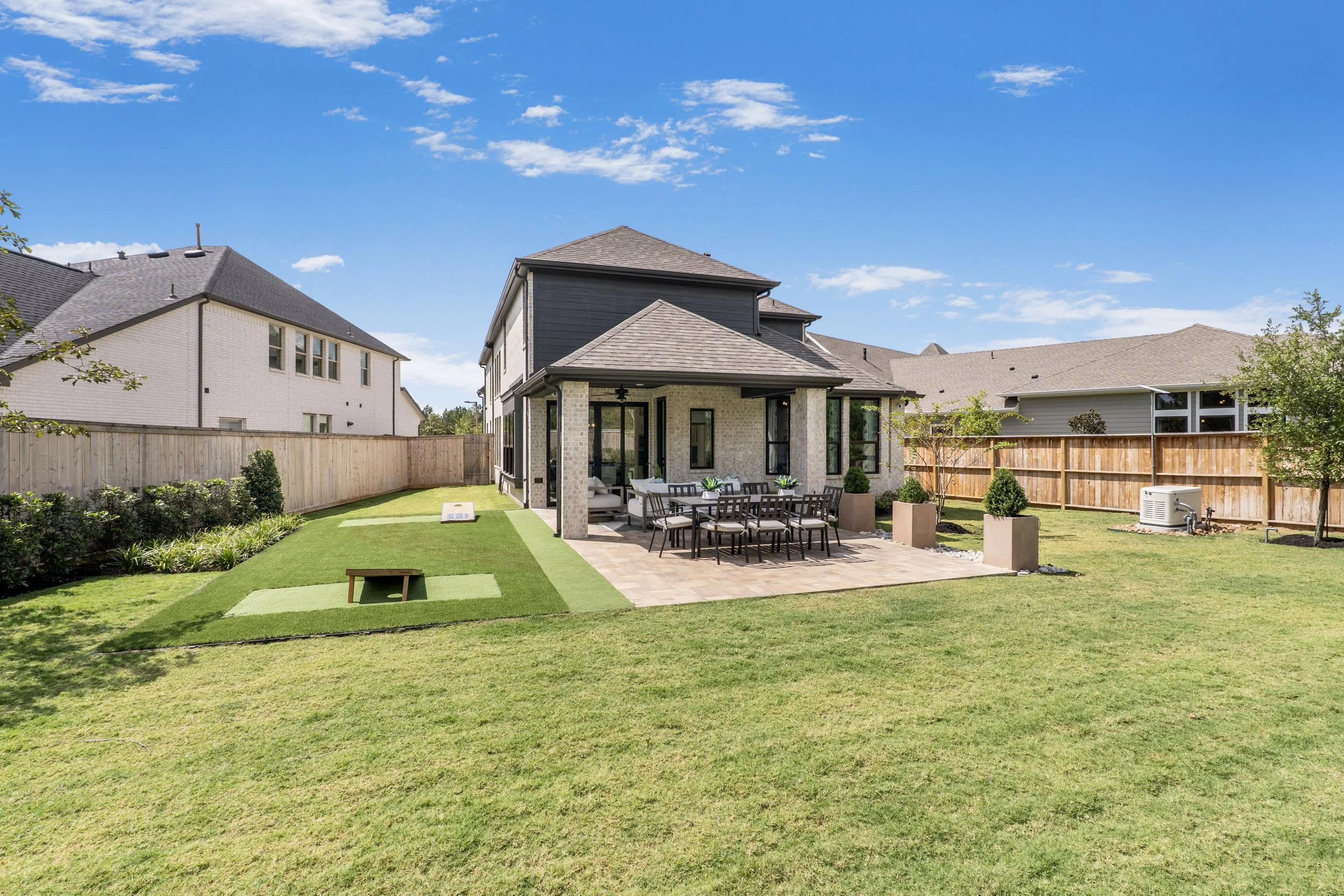 Backyard with a house, patio, outdoor dining table, chairs, benches, lawn, and plants under a blue sky.