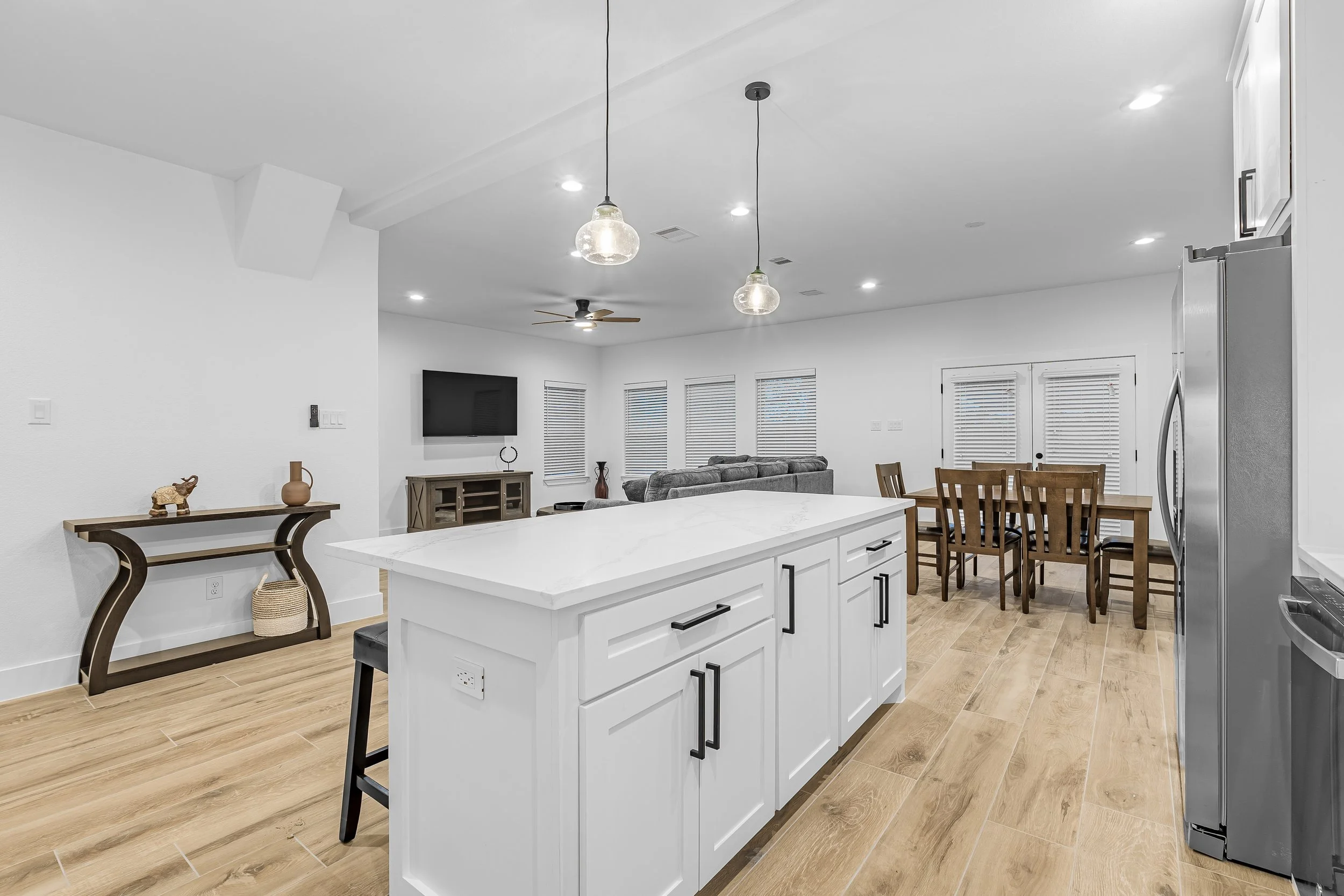 Open-concept living area with kitchen island, dining table, and living room with gray sofa, TV, and windows.