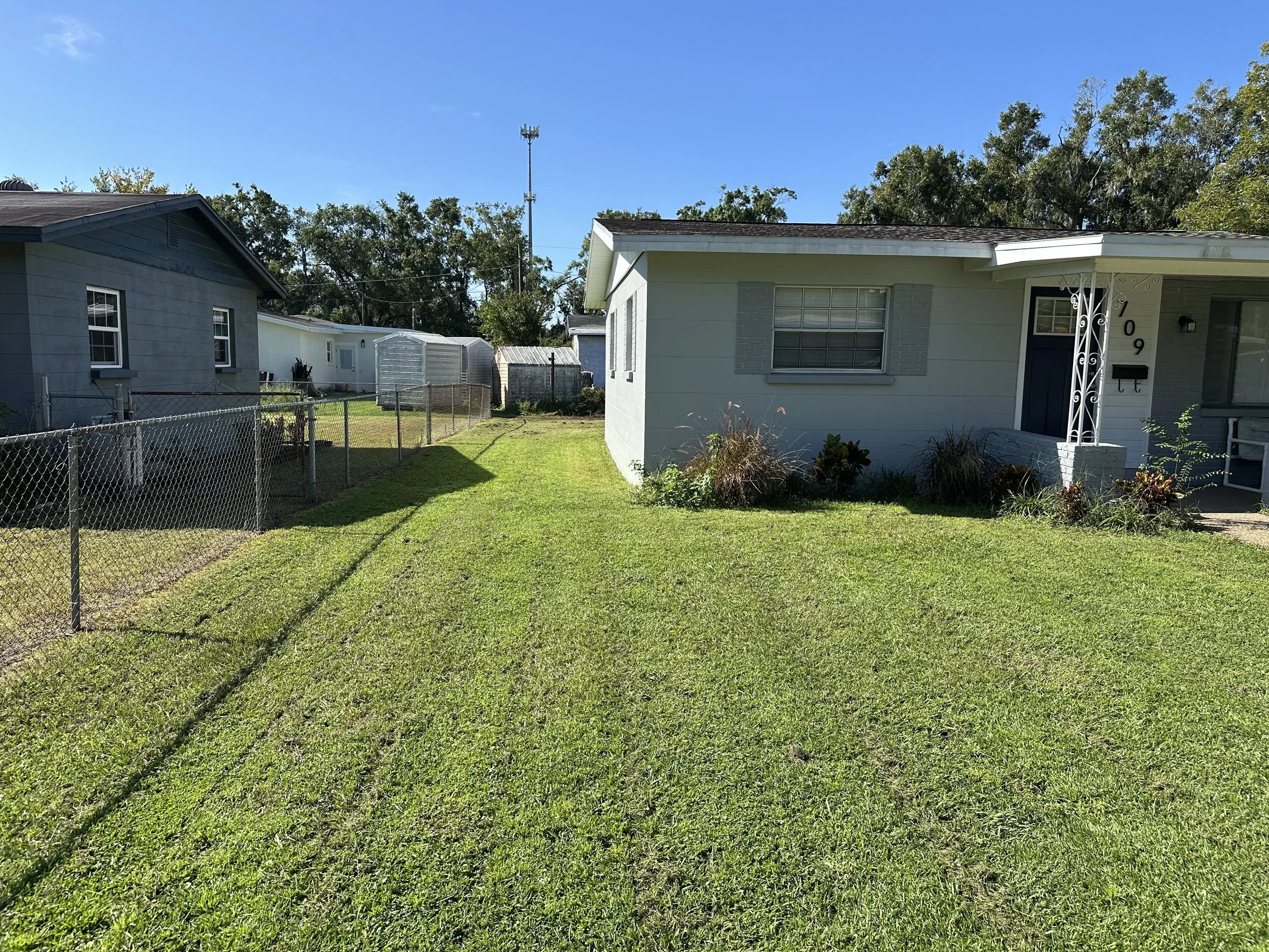 A front yard with green grass, a white house with blue shutters, and a garden bed with plants near the house. Other houses and trees are visible in the background under a clear blue sky.