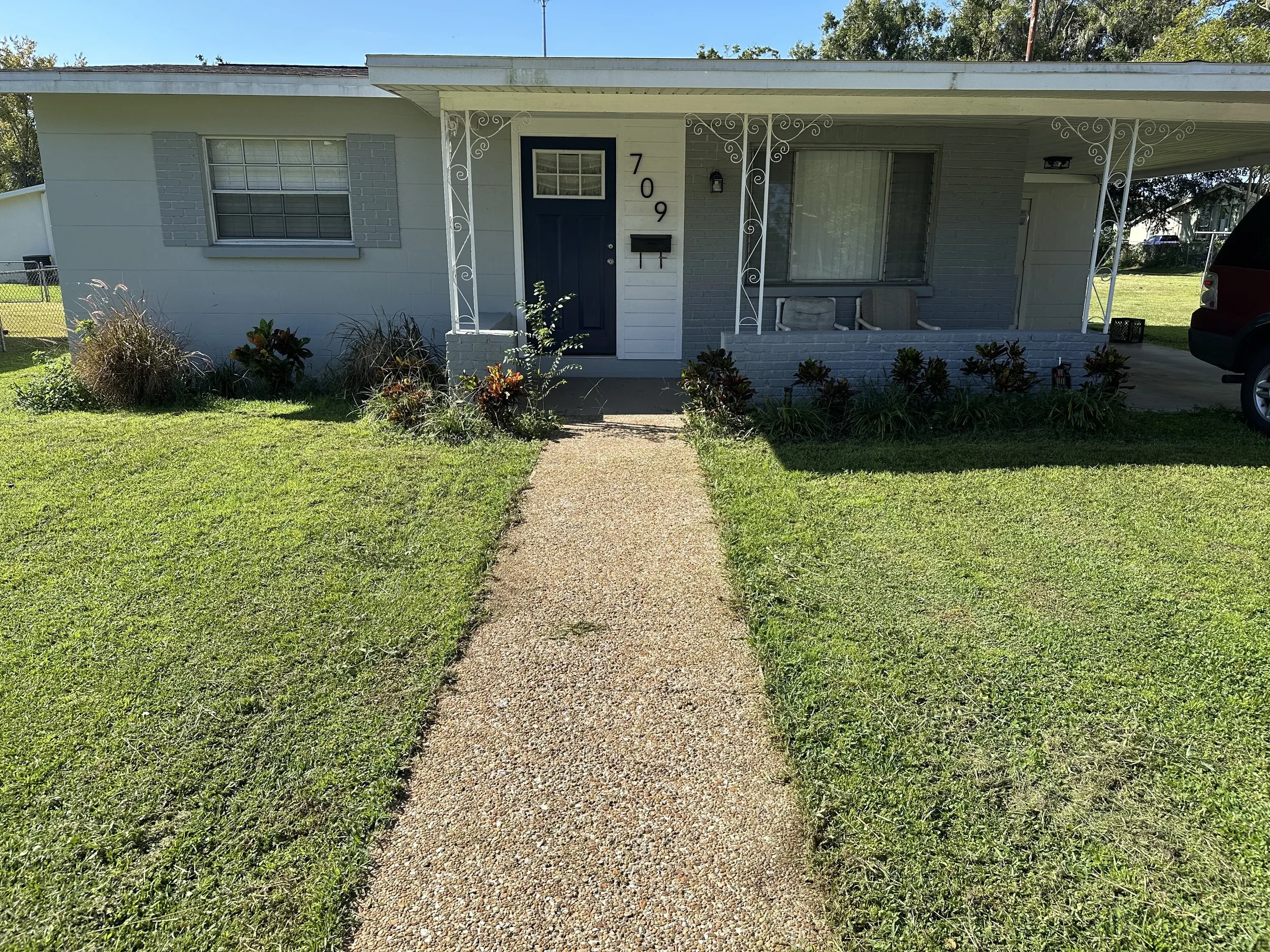 Front view of a single-story house with a concrete sidewalk leading to the front door, white and gray exterior, porch with decorative iron columns, and a small garden with shrubs and flowers.
