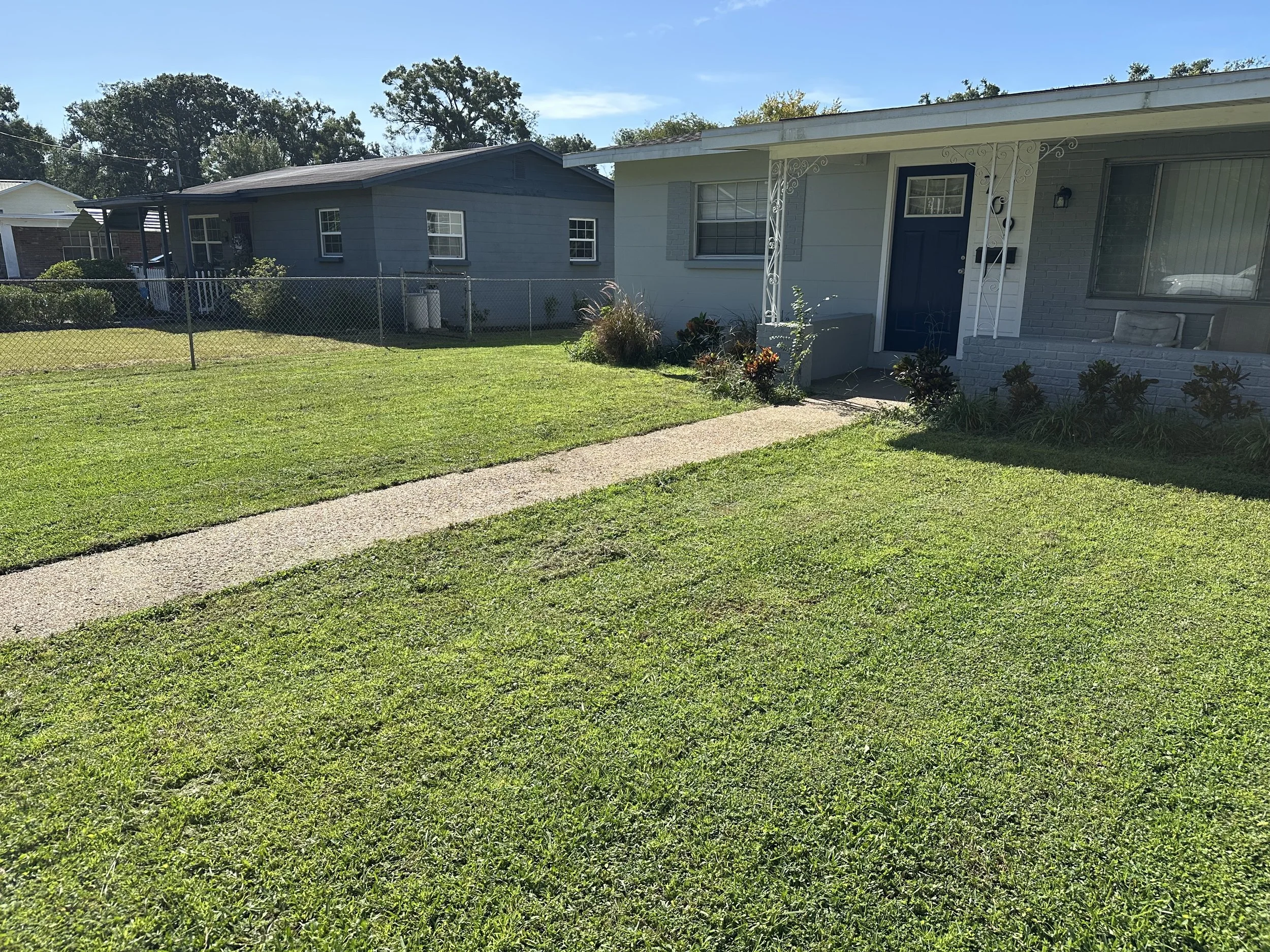 Front yard of a house with a concrete walkway, green lawn, some plants, and a blue front door, with neighboring houses in the background.