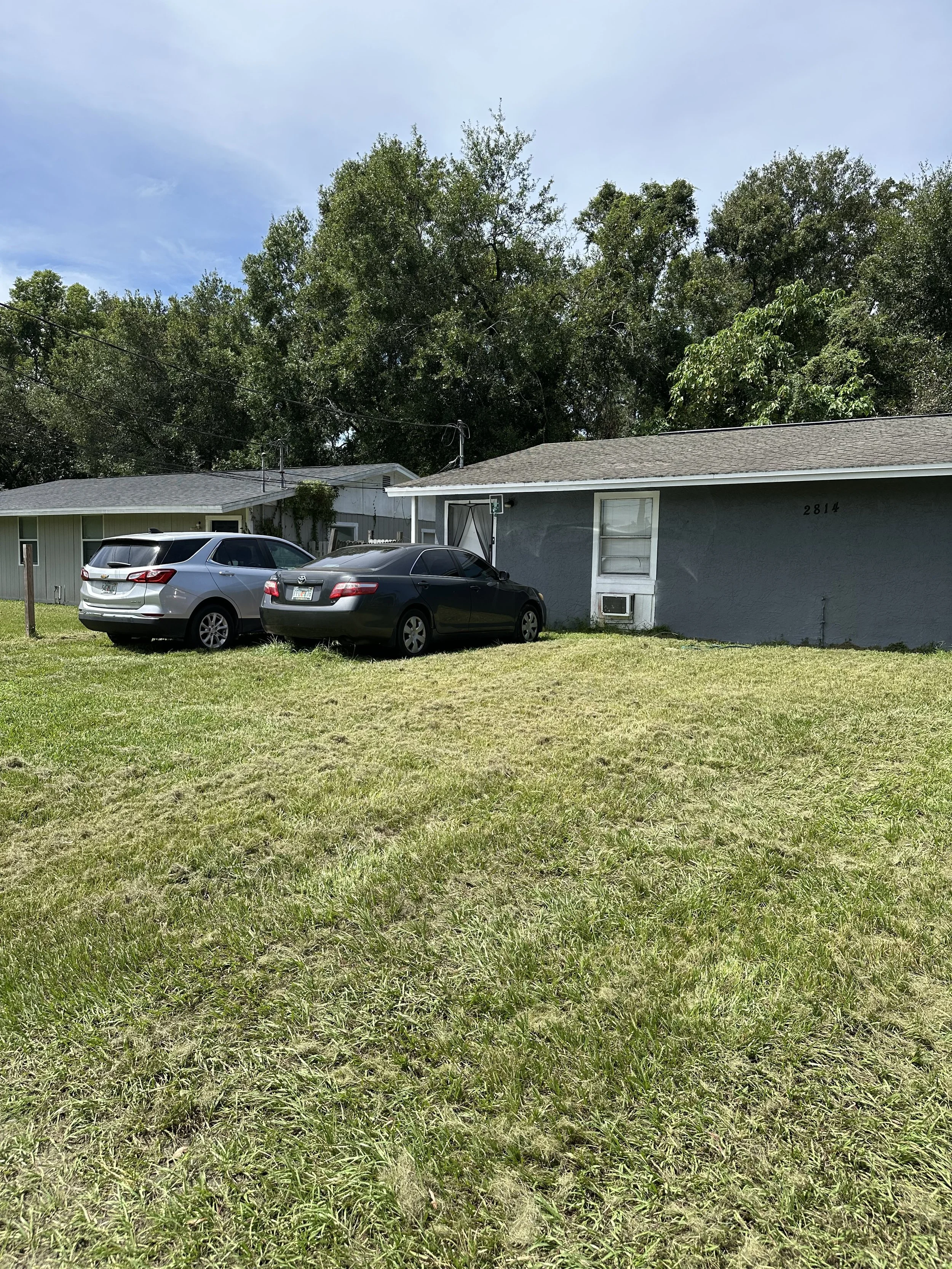 Front yard with grass, two parked cars, one black and one gray, in front of a single-story gray house with window air conditioning unit, against a backdrop of trees and a blue sky.