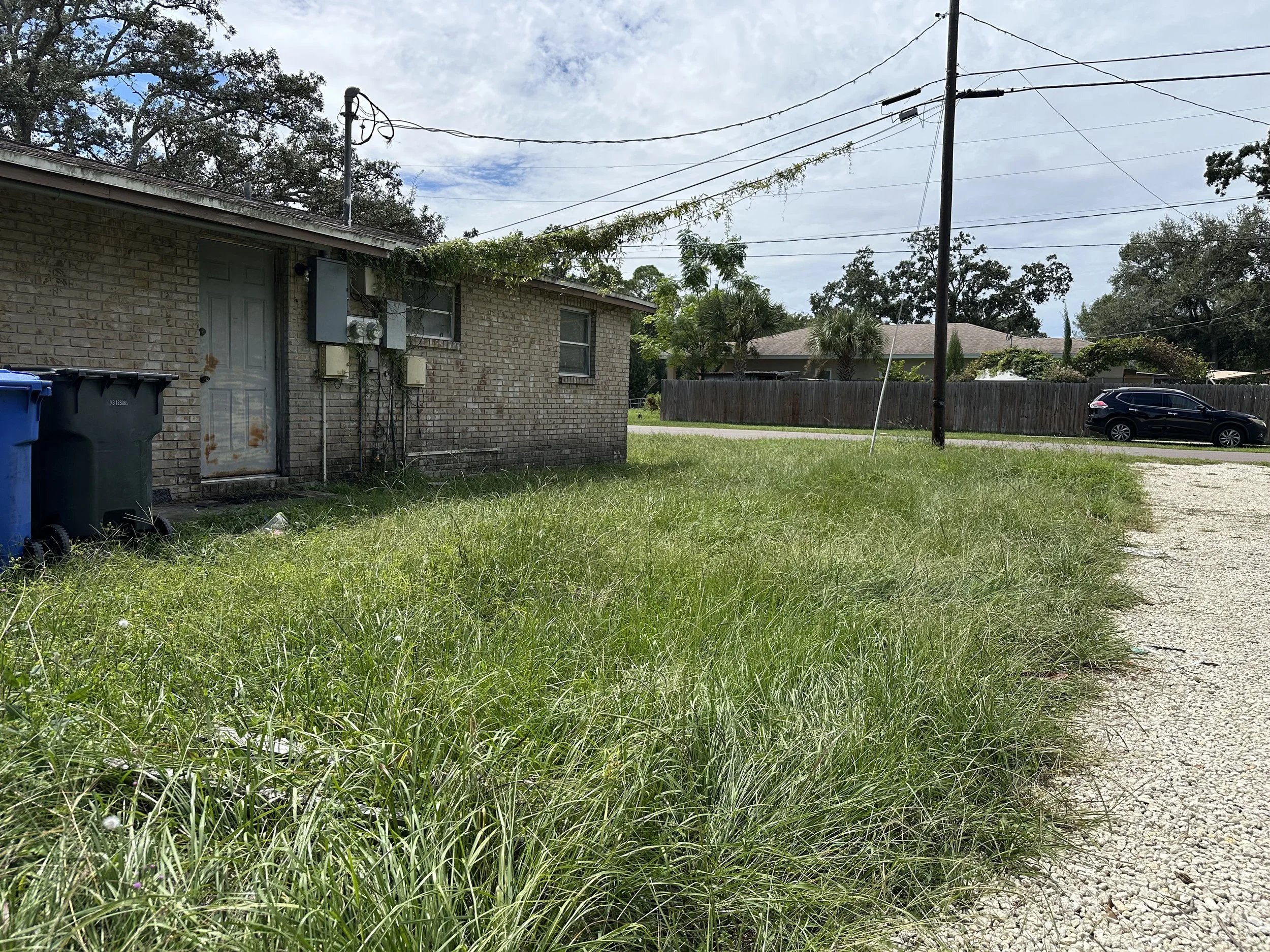 Side view of a house with a brick wall, utility boxes, and overgrown grass in the foreground. Power lines and a parked car are visible in the background under a partly cloudy sky.