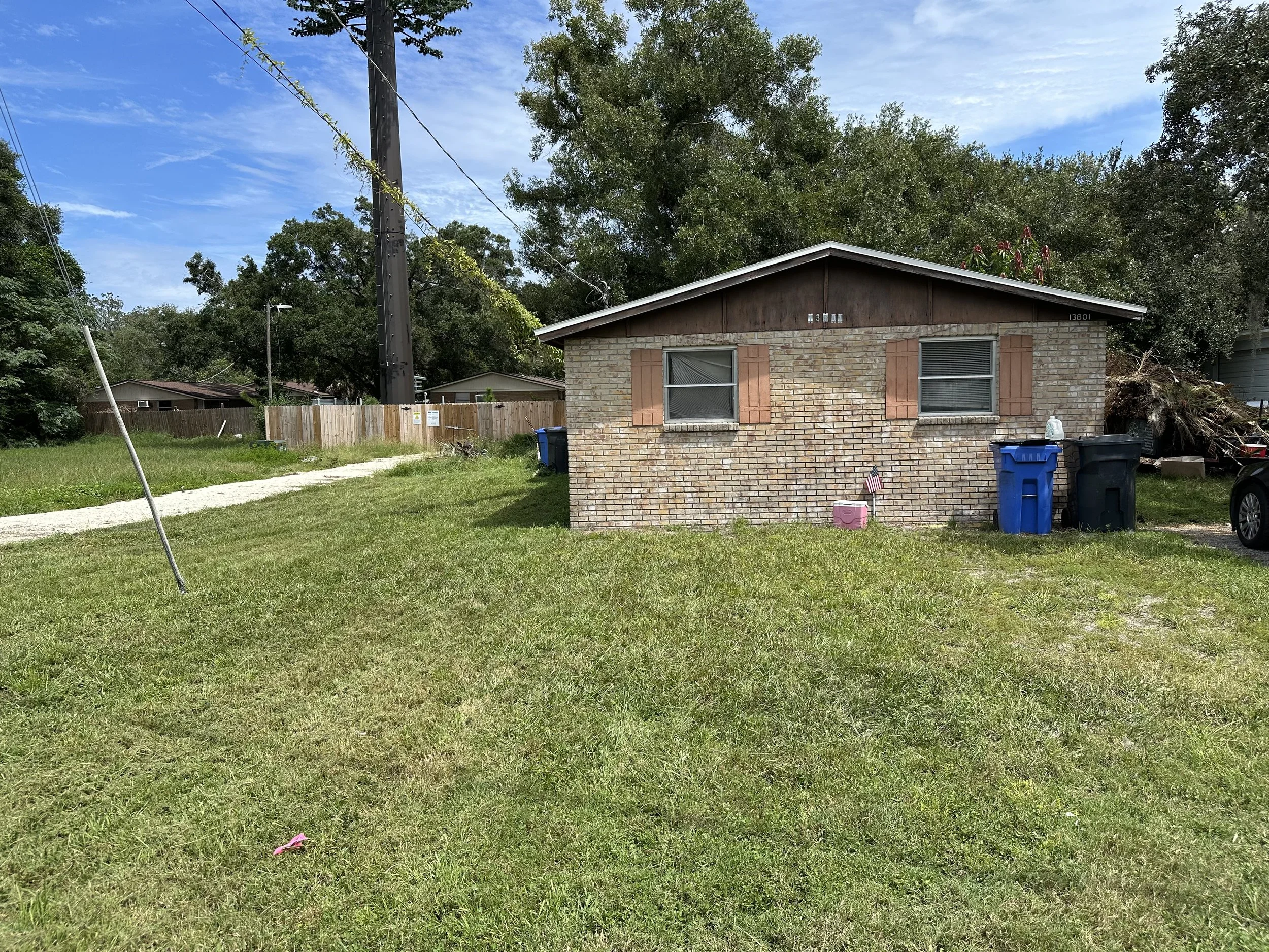 A small brick house with two windows, pink shutters, and a sloped roof. The house has a grassy front yard with trash bins near it, a flag, and a small pink object on the ground. Behind, there are trees, a wooden fence, and a dirt path leading to the
