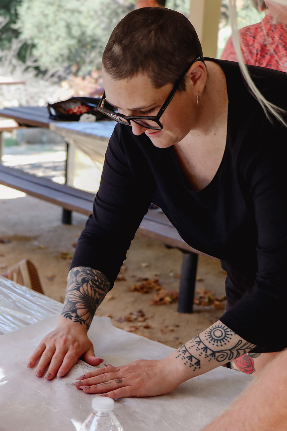 A woman with short hair, tattoos on her arms, and glasses, leaning over a table, working with her fish print at a gyotaku workshop hosted by Jason Bero.