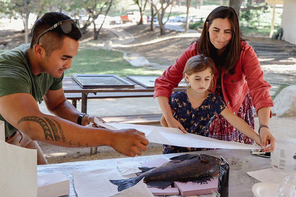 Jason Bero assisting guests at a gyotaku workshop surrounded by trees and outdoor furniture.
