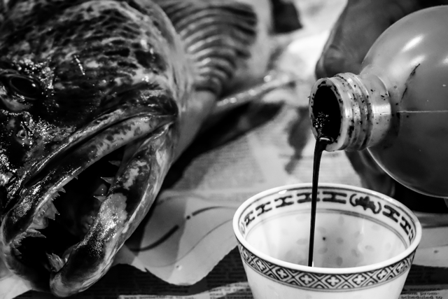 A black-and-white photo of a fish lying on a surface, with a bottle pouring dark liquid into a decorative bowl nearby.