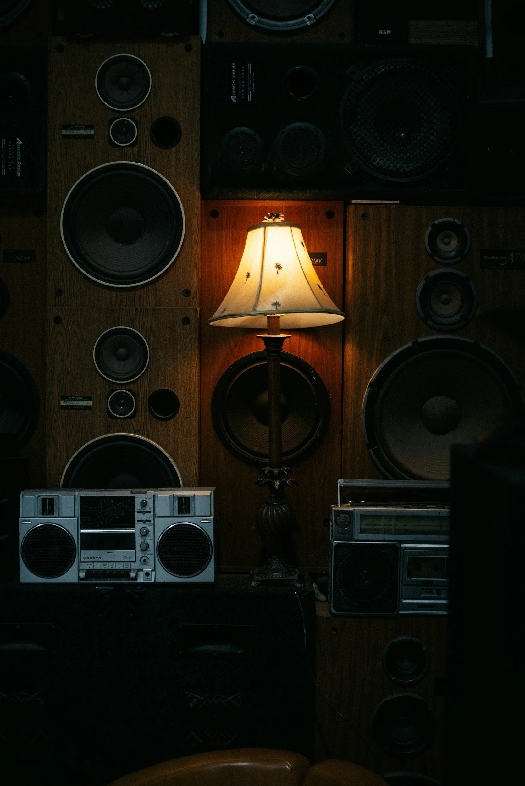 Room with multiple vintage speakers on wooden panels, a floor lamp with a palm tree pattern, and retro stereo systems.