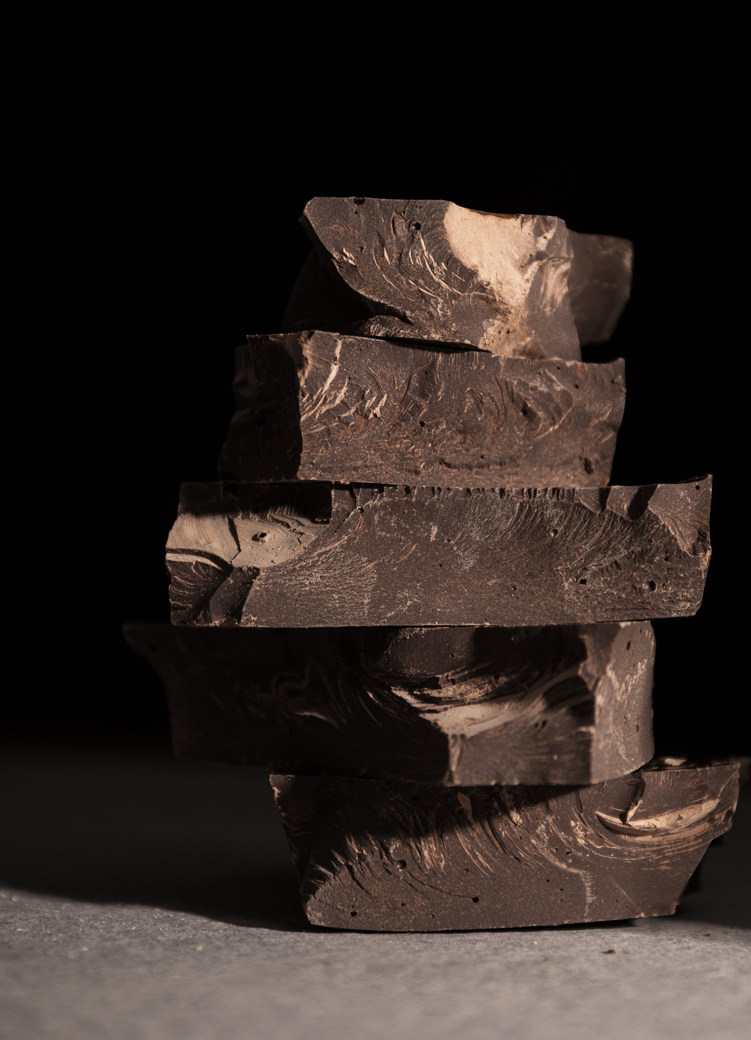 A stack of dark chocolate blocks sits dramatically against a black background, their rough, organic textures highlighted by warm, directional light. Deep shadows and earthy brown tones give the image a **moody, luxurious and slightly mysterious atmos