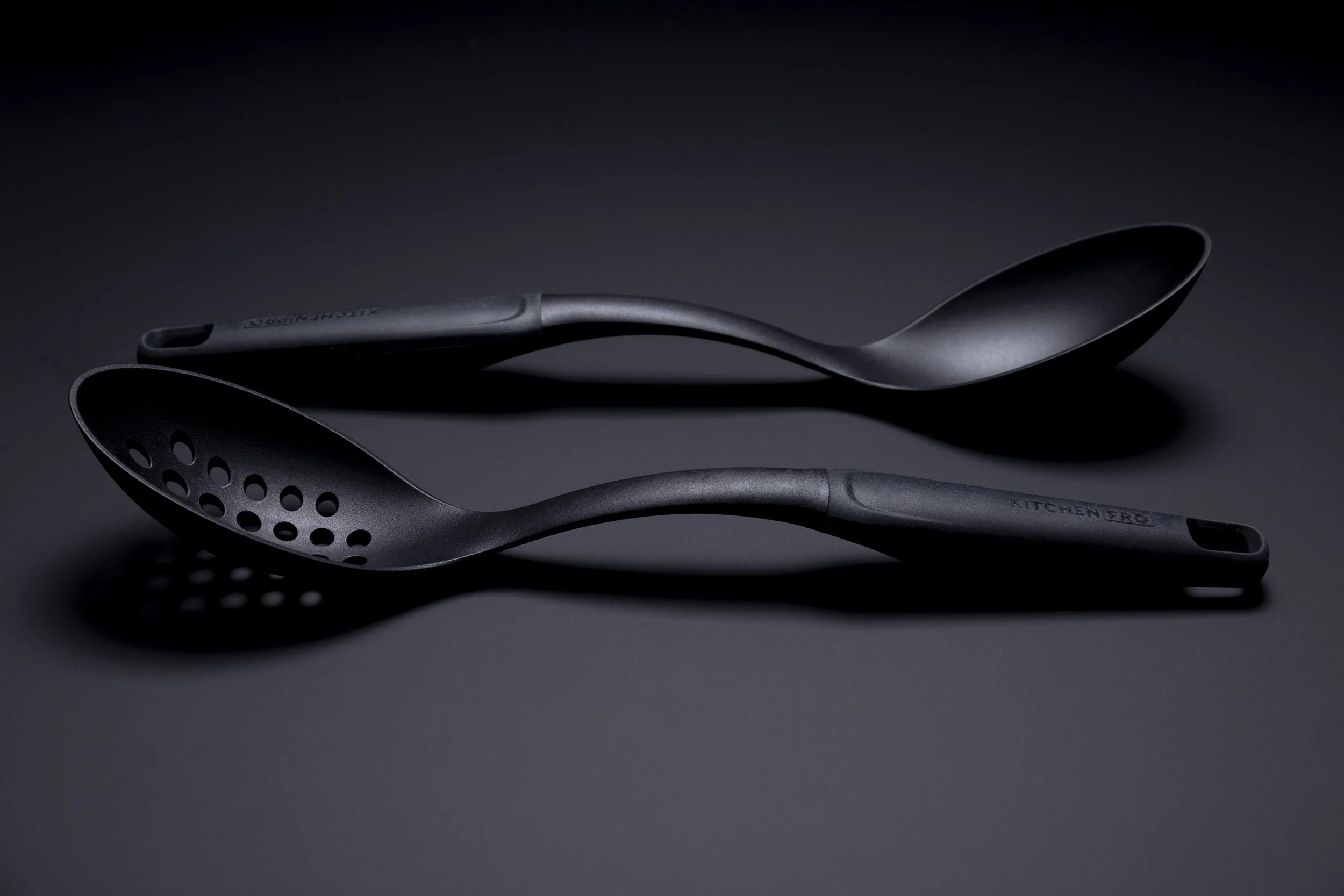 A moody, low-lit photograph showcasing two sleek black kitchen utensils resting side by side. Their smooth, matte finish and gently curved handles are highlighted by soft, directional lighting, creating subtle shadows and a refined, modern aesthetic 