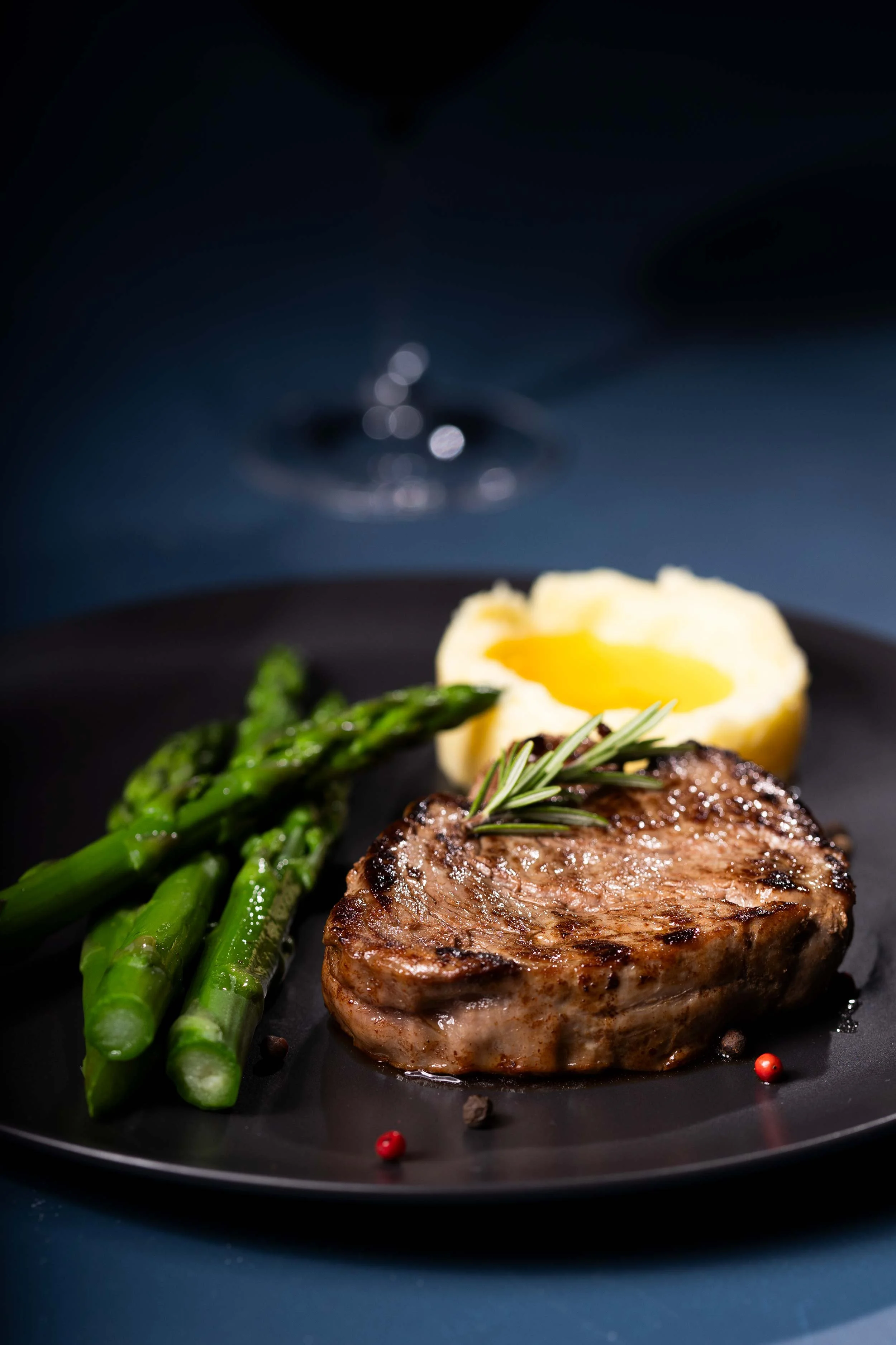 A perfectly seared rare steak rests at the centre of the plate, its tender, rosy interior glistening with juices. Alongside, vibrant asparagus spears add a fresh, green crunch, while a cloud of smooth, buttery mashed potato completes the dish with co