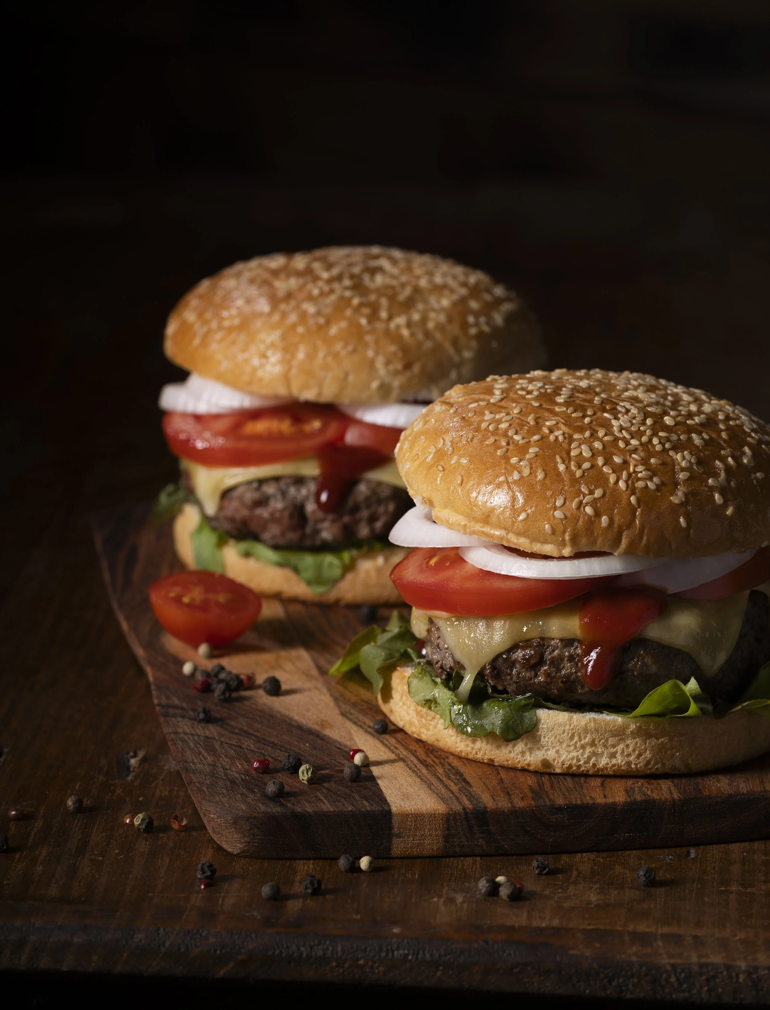 A dark, atmospheric shot captures two stacked burgers resting side by side, their glossy buns catching a soft, low light. Shadows deepen the textures of melted cheese and charred edges, giving the scene a rich, indulgent mood.