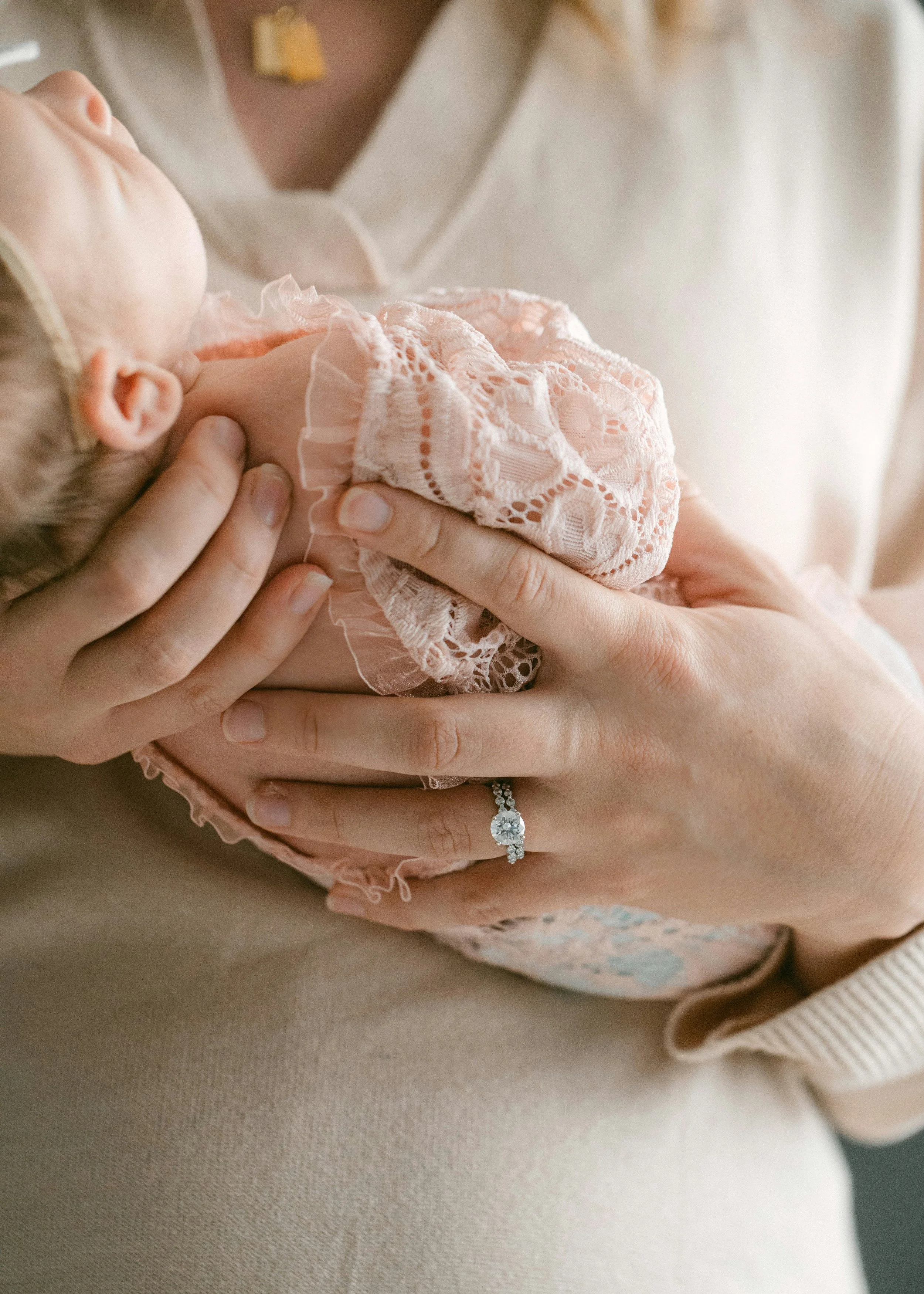 Close-up of a woman holding a newborn baby, with the woman's hand showing an engagement ring and delicate clothing covering her chest.