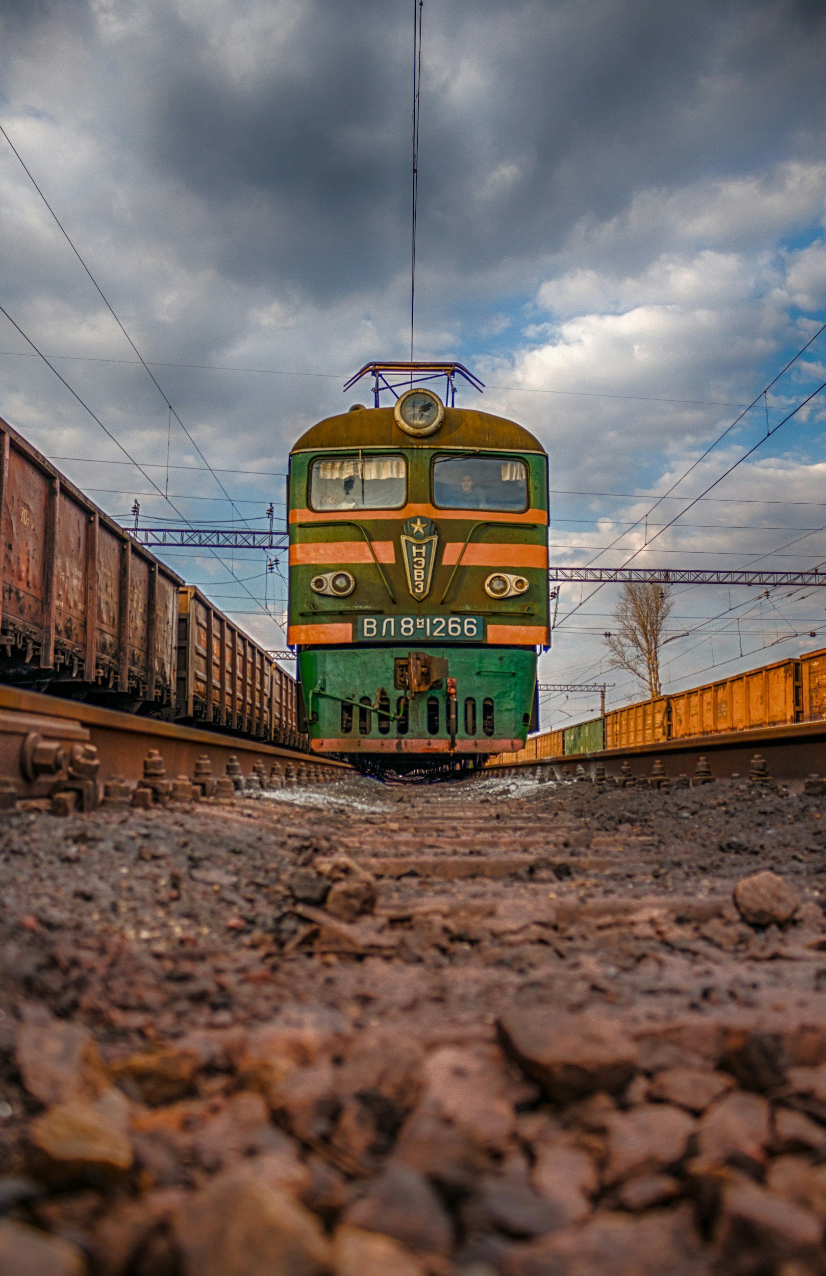 Low-angle view of a vintage green and orange electric train approaching on railway tracks, with freight cars on both sides and a cloudy sky overhead.