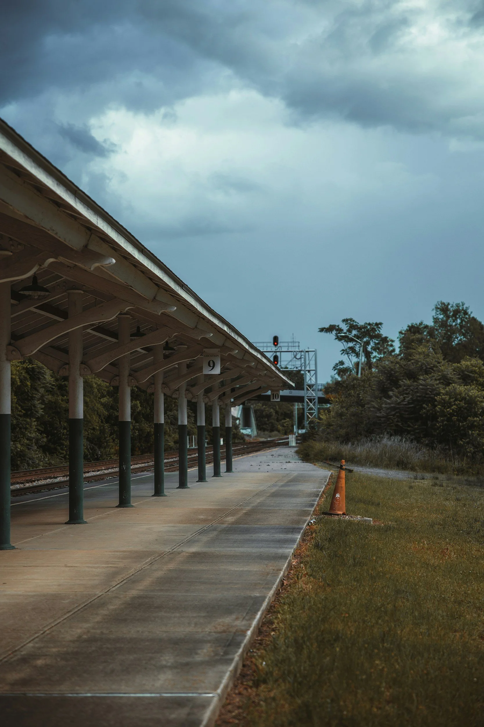 Empty train station platform with a canopy, traffic signal showing red, and surrounding greenery, under a cloudy sky.