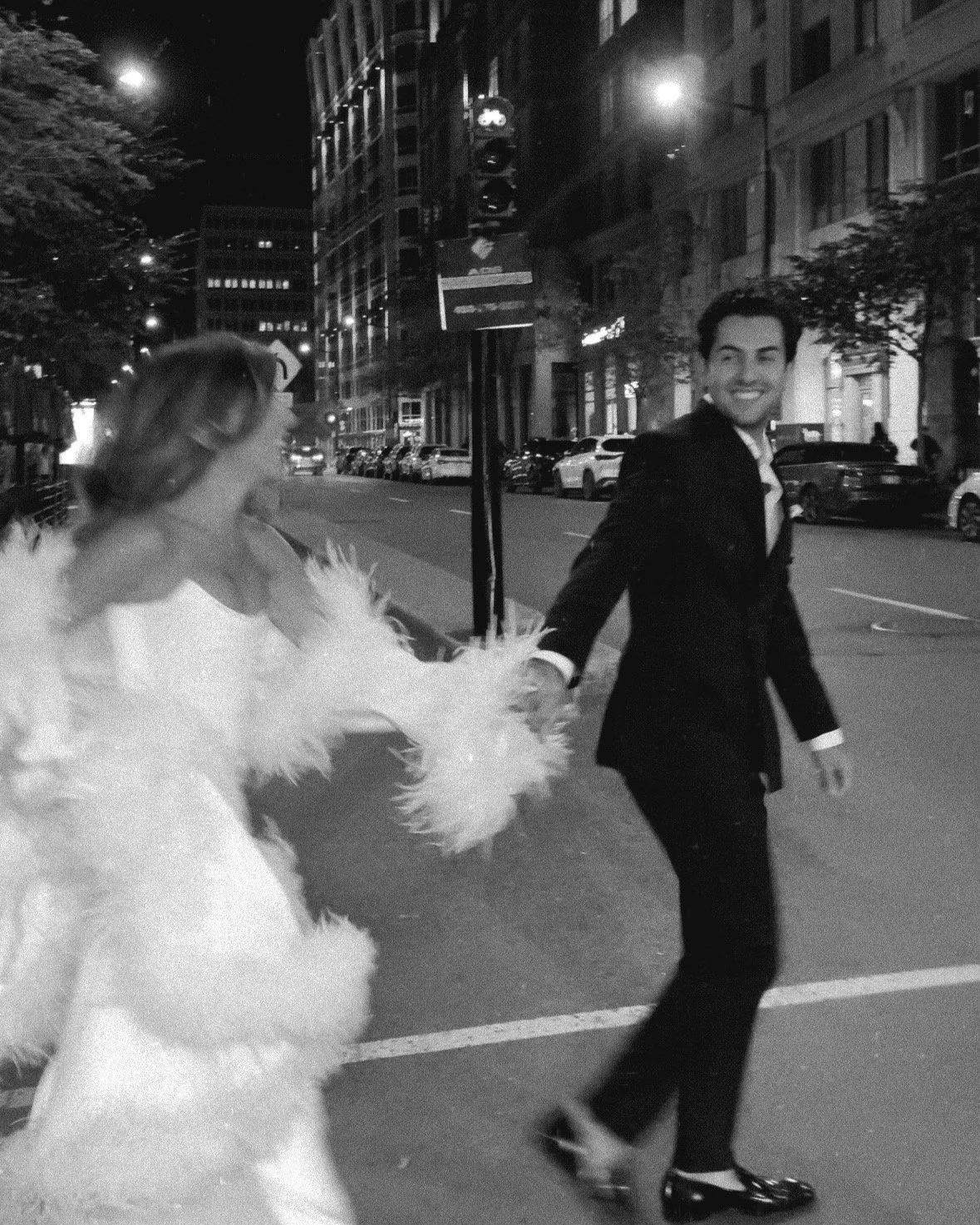 A man and a woman walking hand-in-hand on a city street at night. The woman is dressed in a white gown with feathered details, and the man is in a black tuxedo. The scene is illuminated by streetlights, and cars are parked along the street.