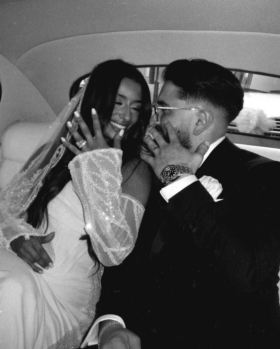 A happy couple on their wedding day, sitting inside a car, sharing an intimate moment. The bride is smiling and wearing a veil and wedding dress, while the groom, dressed in a tuxedo, is gently holding her face.