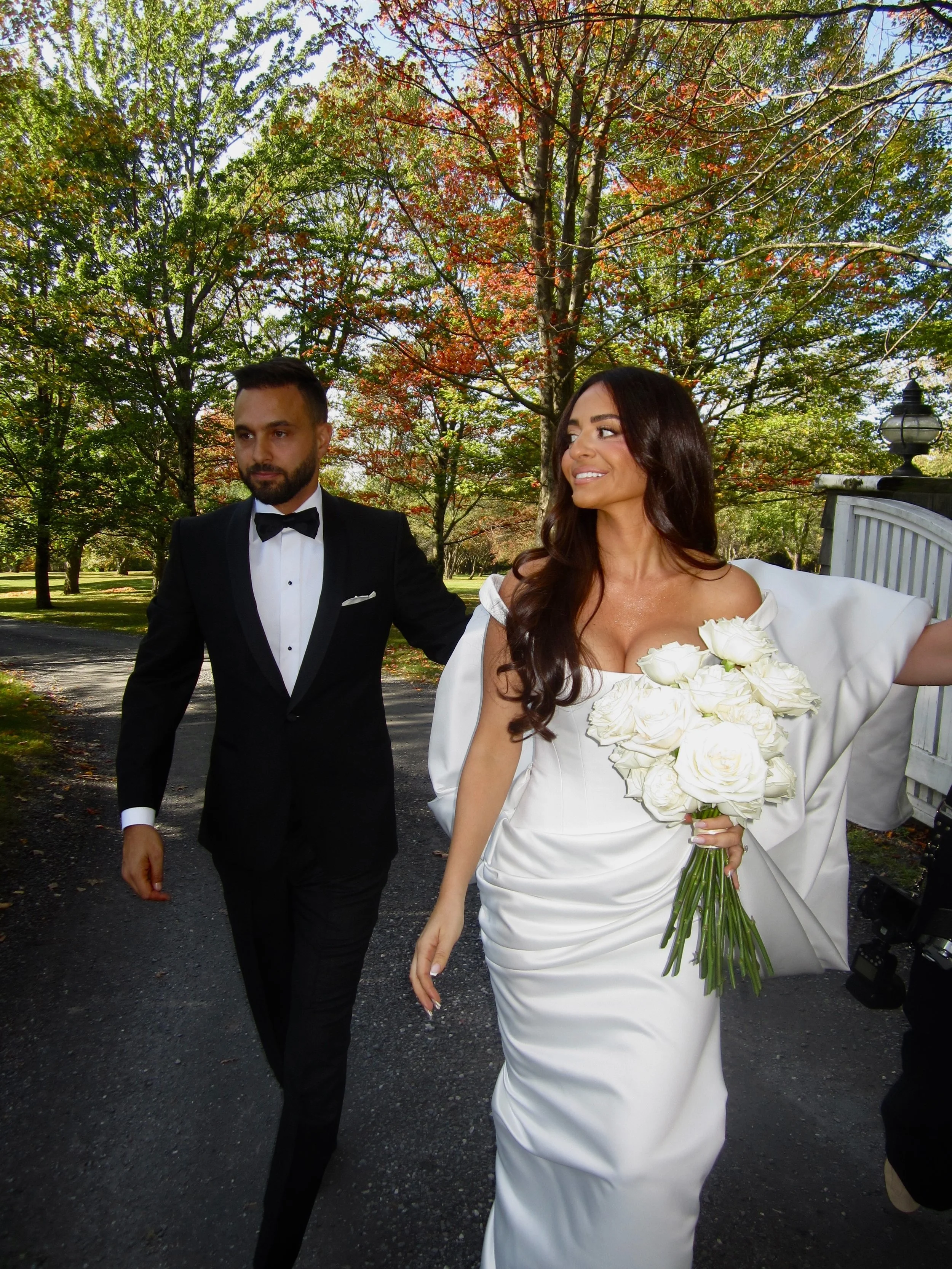 A bride in a white wedding dress holding a bouquet of white roses walking with a groom in a black tuxedo outdoors in a park with autumn trees.