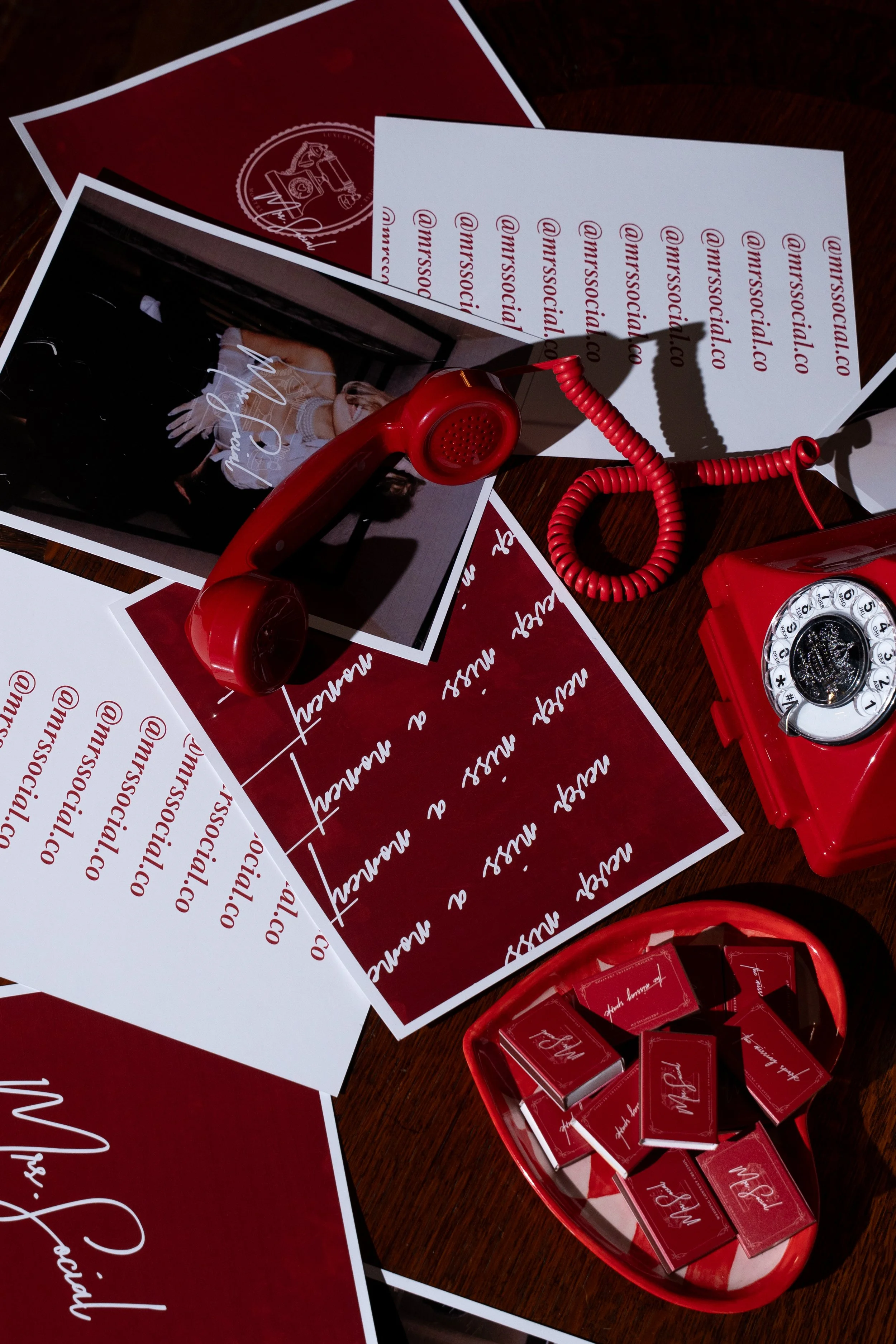 A red rotary telephone, printed sheets with social media handle @misssocalico, a magazine photo of a woman, a bowl with small red and black boxes, and the magazine logo on a dark wood surface.