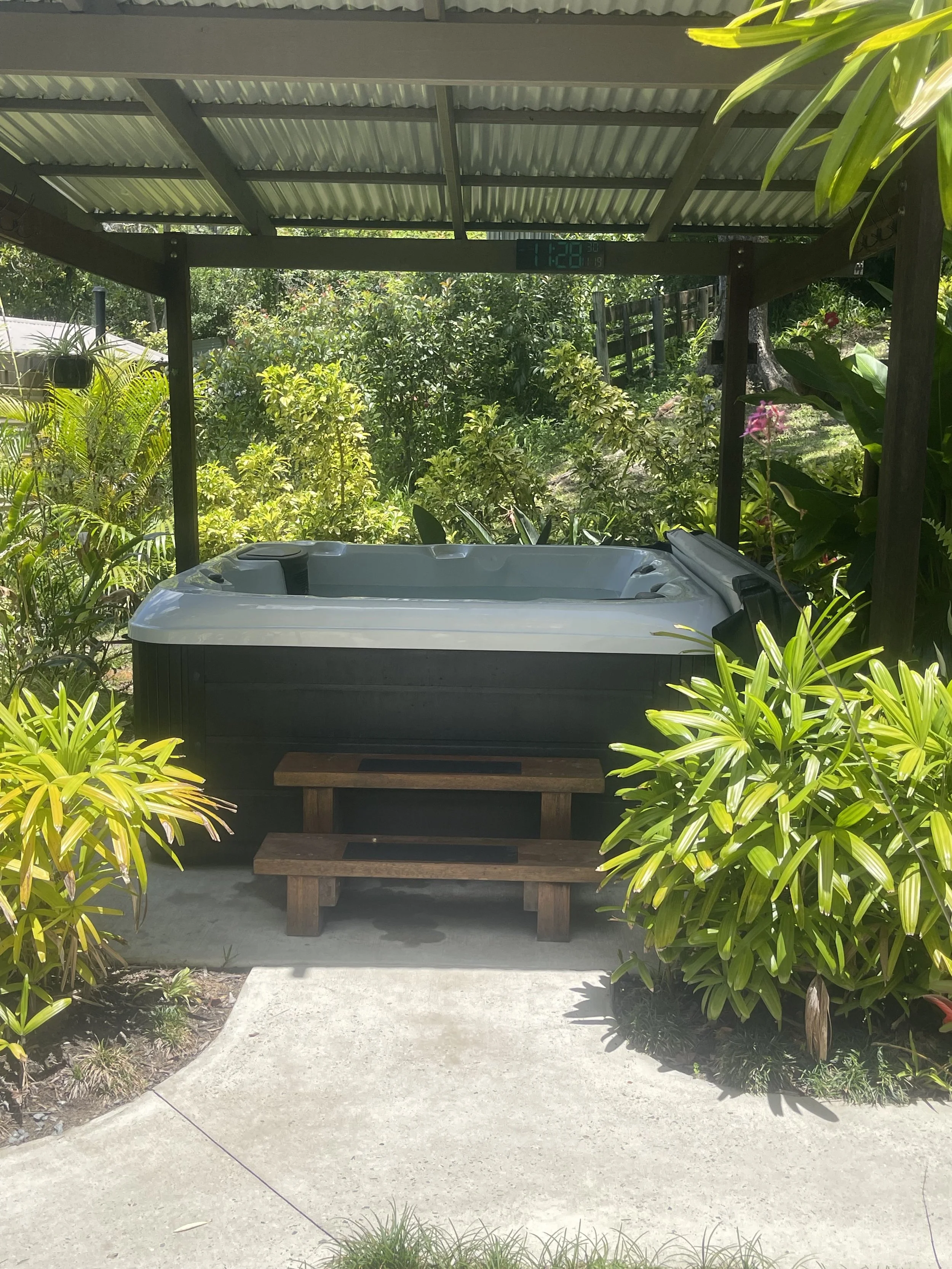 An outdoor hot tub or jacuzzi situated on a concrete patio beneath a corrugated metal roof, surrounded by lush green plants and garden foliage.