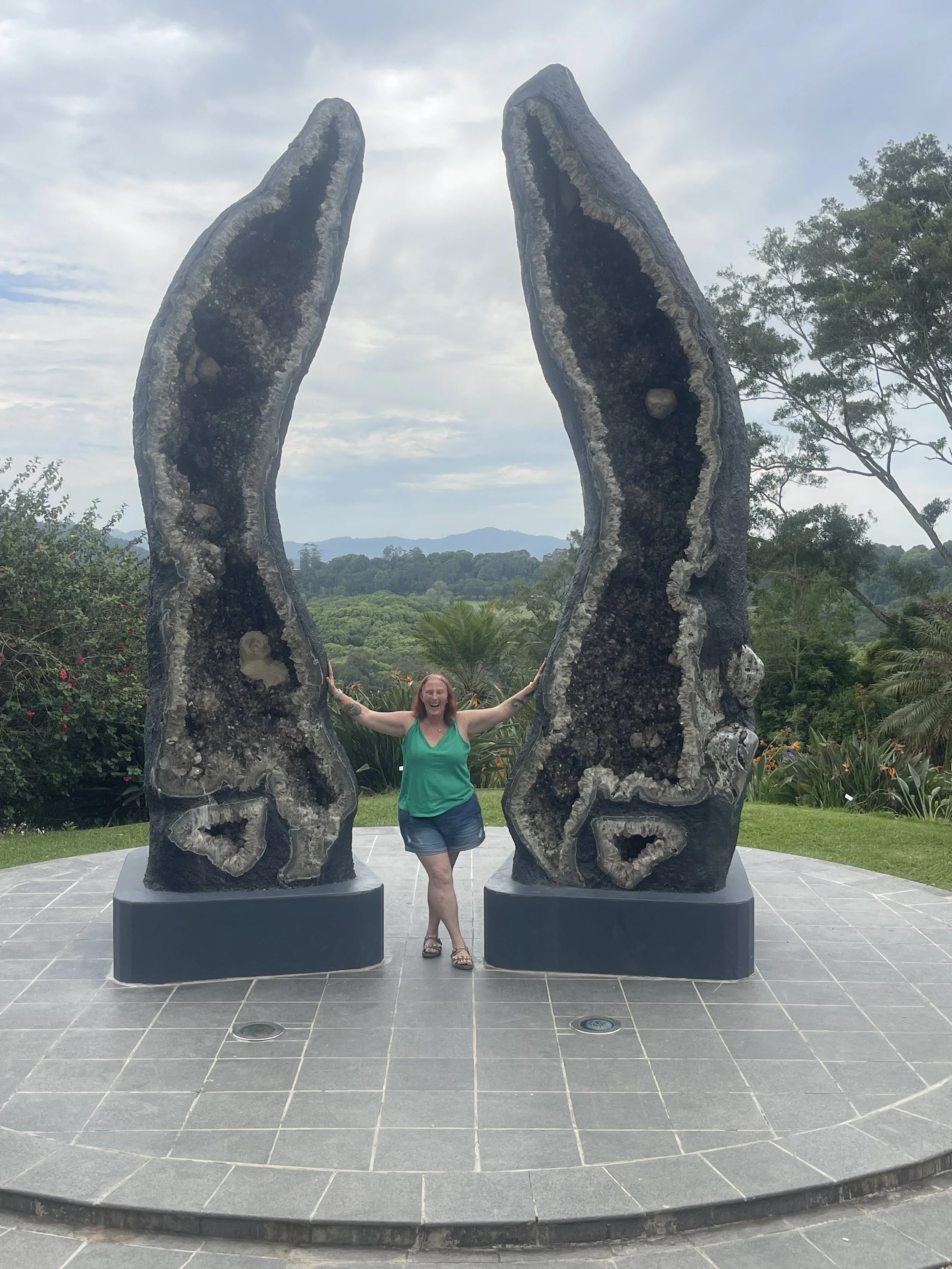 A woman in a green top and shorts standing between two large geode sculptures on a stone platform outdoors with trees and mountains in the background.