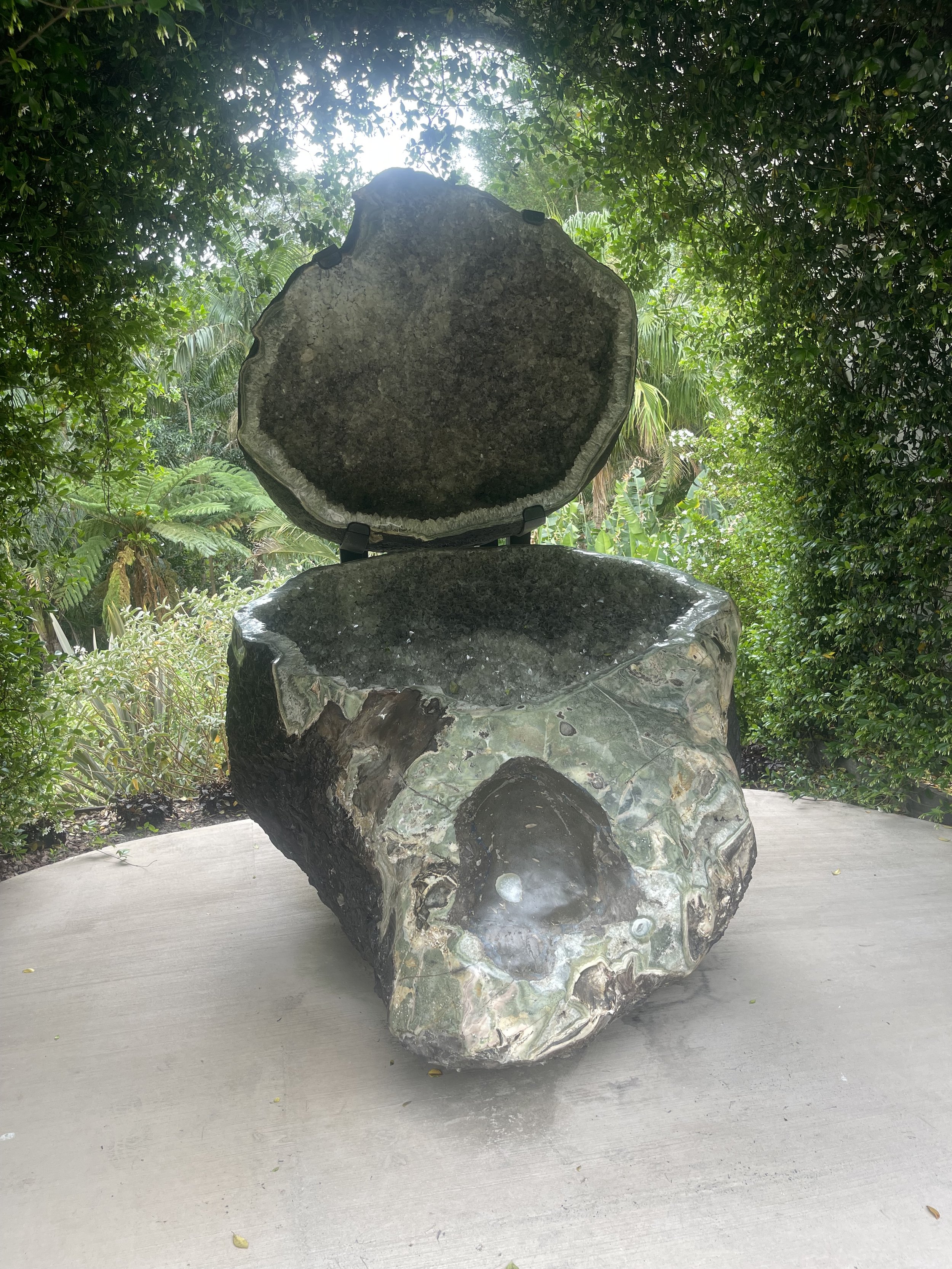 A polished geode with an opening on top, placed on a wooden surface outdoors surrounded by green foliage.