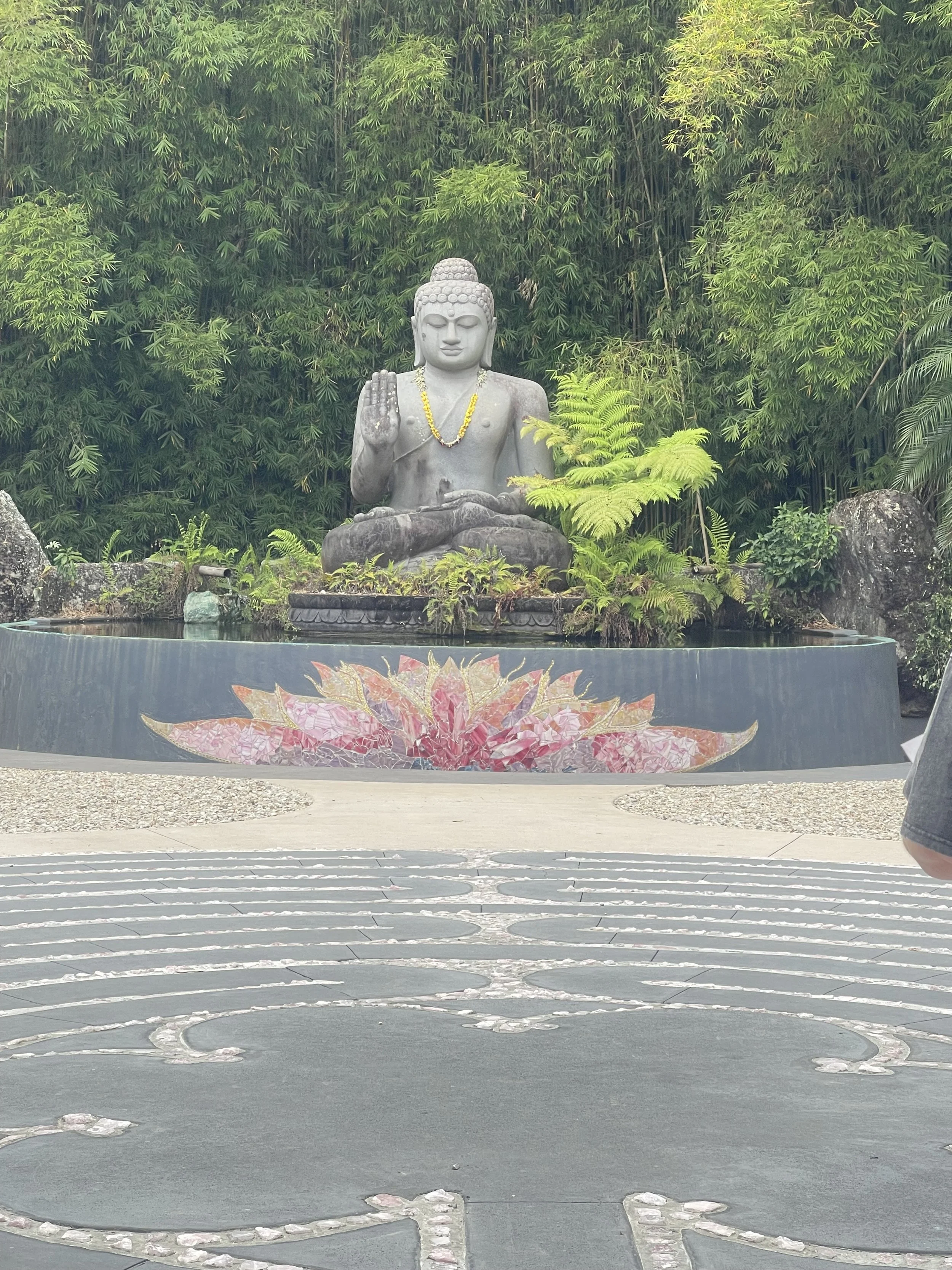 A large Buddha statue sitting in a meditation pose in front of lush green foliage, with a decorative water fountain at its base featuring a pink lotus design.