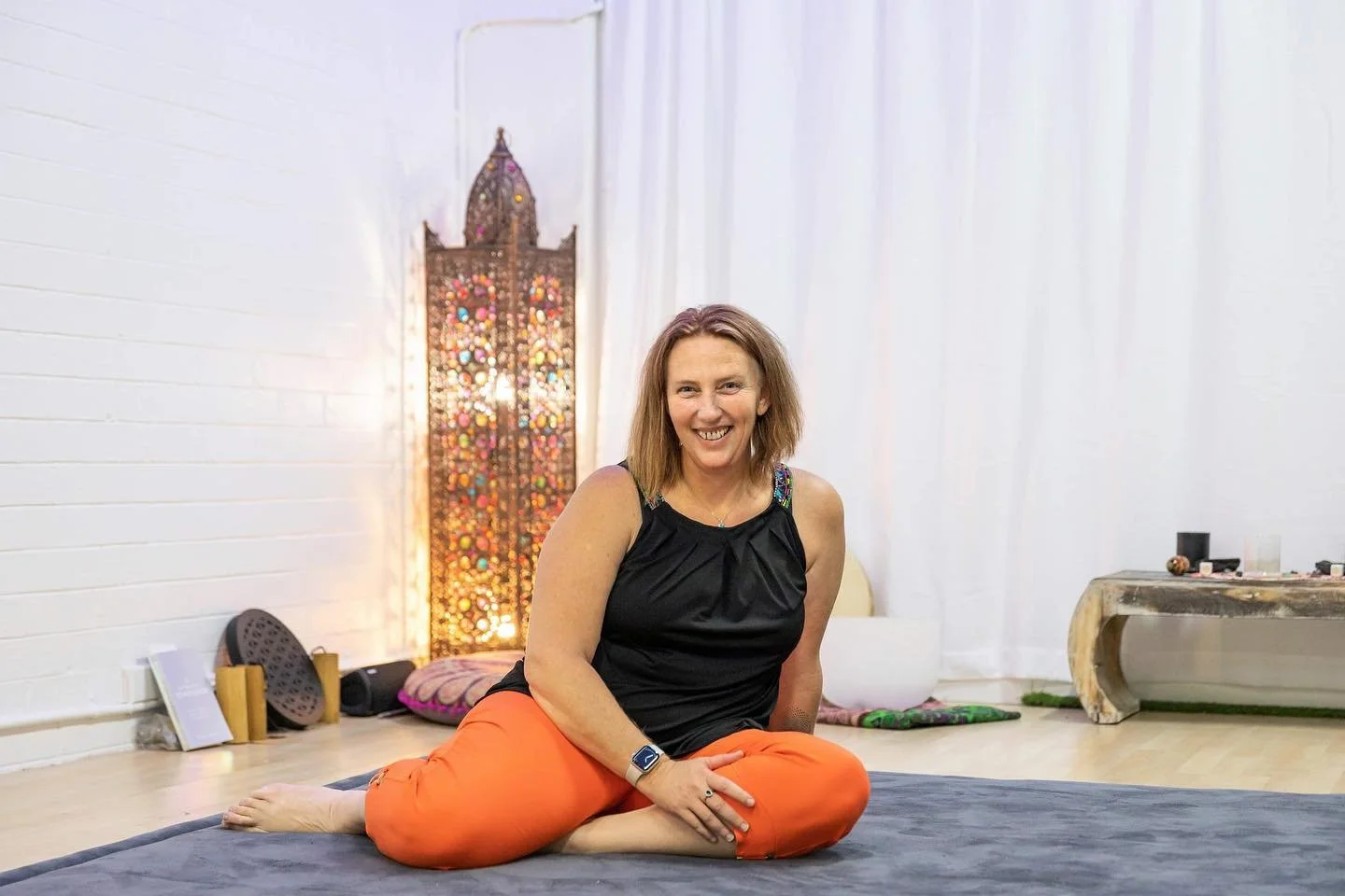 Woman sitting on yoga mat, smiling with legs crossed, in a room with white walls, a decorative lantern in the background, and yoga props on the floor.