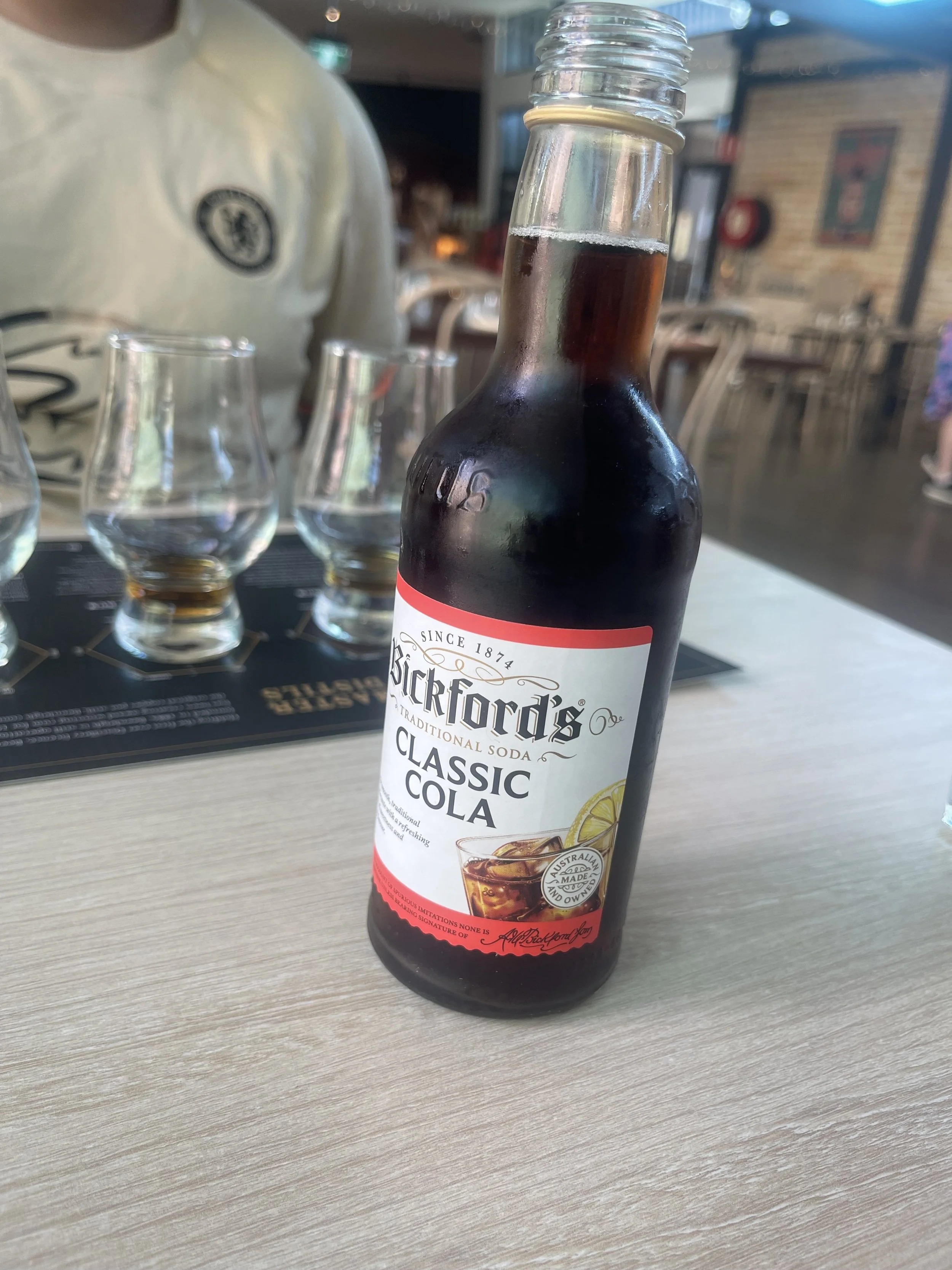 A bottle of Bickford's classic cola soda on a table in a restaurant, with empty glasses and a person in the background.