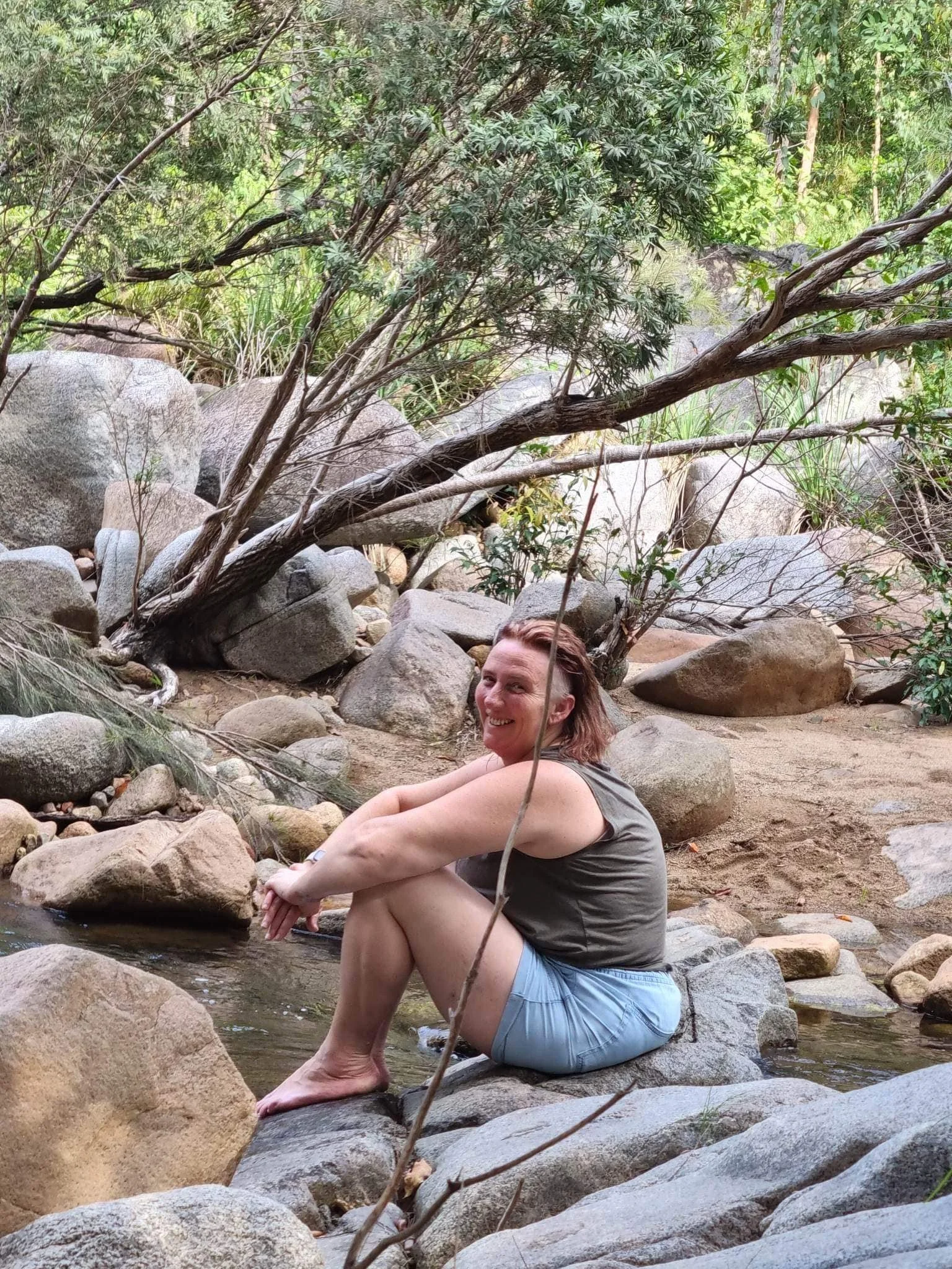 A woman sitting on rocks in a creek, smiling, surrounded by trees and greenery.