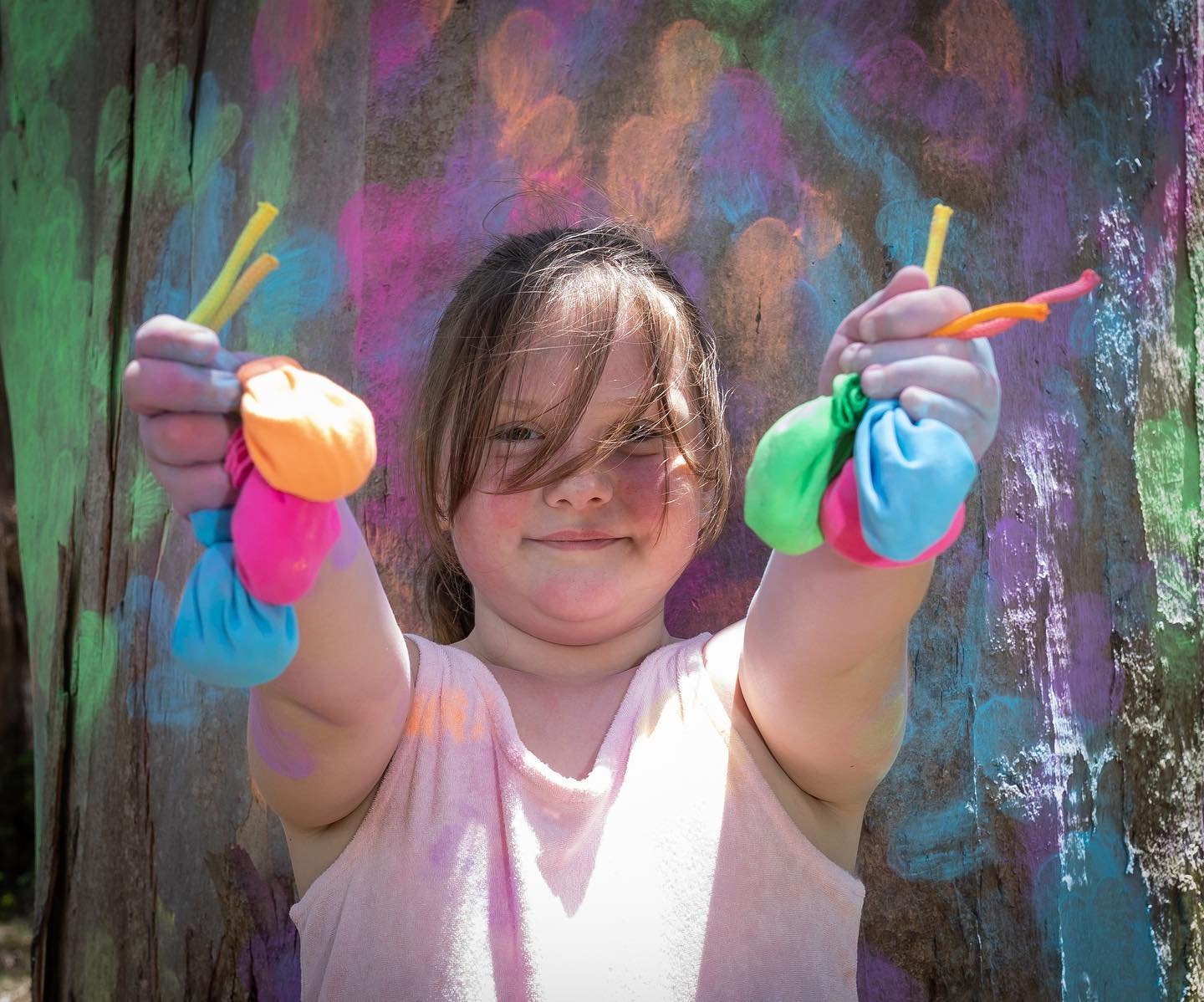 Young girl holding colorful paint-covered cloth bags and paintbrushes in front of a vibrant, chalk-covered wall.